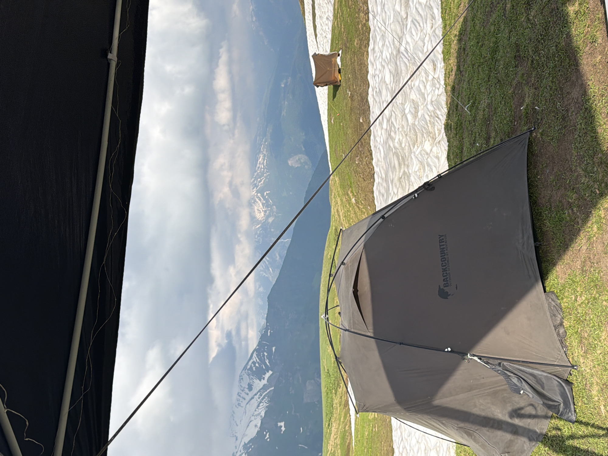 An overhanging black canopy casts a shadow over the foreground of the image, framing a view of a mountainous landscape. A dark gray tent, labeled with the "BACKCOUNTRY" logo, is pitched on a grassy slope dotted with patches of melting snow. In the mid-ground, another smaller, brown shelter is visible on a similar slope. The background is dominated by a range of snow-capped mountains under a cloudy sky, suggesting a high-altitude environment. The scene depicts a campsite in the vicinity of Mestia, Georgia, a region known for its dramatic mountain scenery and opportunities for outdoor adventure.
