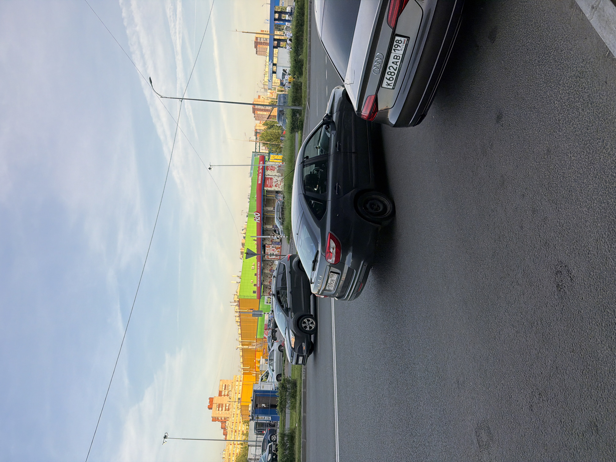 The image captures a street scene in Saint Petersburg, Russia, likely during the late afternoon or early evening, given the warm lighting. The foreground features the rear of a dark grey Audi vehicle with a Russian license plate reading "K682AB 198". To its left and slightly behind is a dark grey Lada car, its rear also visible, with a license plate that appears to read "K109OB 198". In the middle ground, a dark grey SUV, possibly a Ford, is in motion, with its rear right quarter visible. Further back, a light silver sedan is also visible. The background is dominated by a row of modern, brightly colored buildings, with facades in yellow, orange, and green, housing commercial establishments, indicated by signs like "H2O" and "МОЙКА 24 ЧАСА" (Car Wash 24 Hours). Tall streetlights and electrical wires crisscross the sky. The sky is a pale blue with wispy clouds. The street is paved with asphalt, and there is a sidewalk or median with green bushes. The overall impression is of a typical urban environment with ongoing traffic and commercial activity.