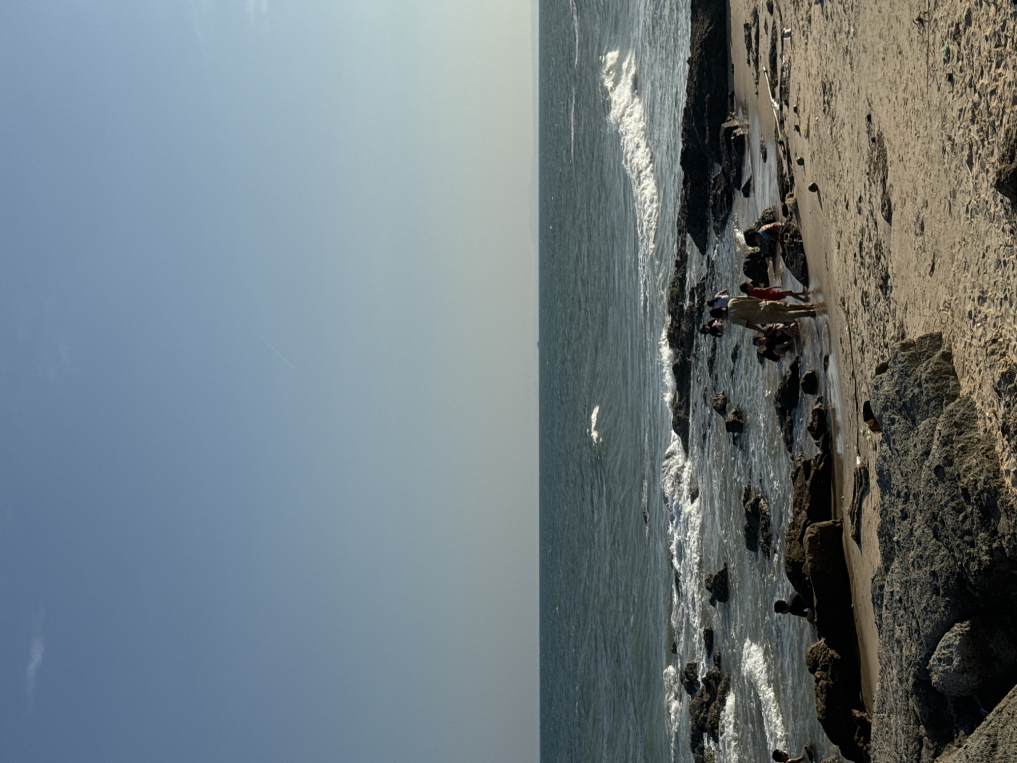 The image depicts a beach scene in Mũi Né, Vietnam. The scene consists of a clear, light blue sky that transitions into a greyish band above the ocean. The ocean has choppy waters and white foam along the shoreline. The beach is composed of light-colored sand with a scattering of rocks and dark, jagged boulders along the coastline. A group of people are standing near the water’s edge, some wearing red. They appear to be interacting with each other.