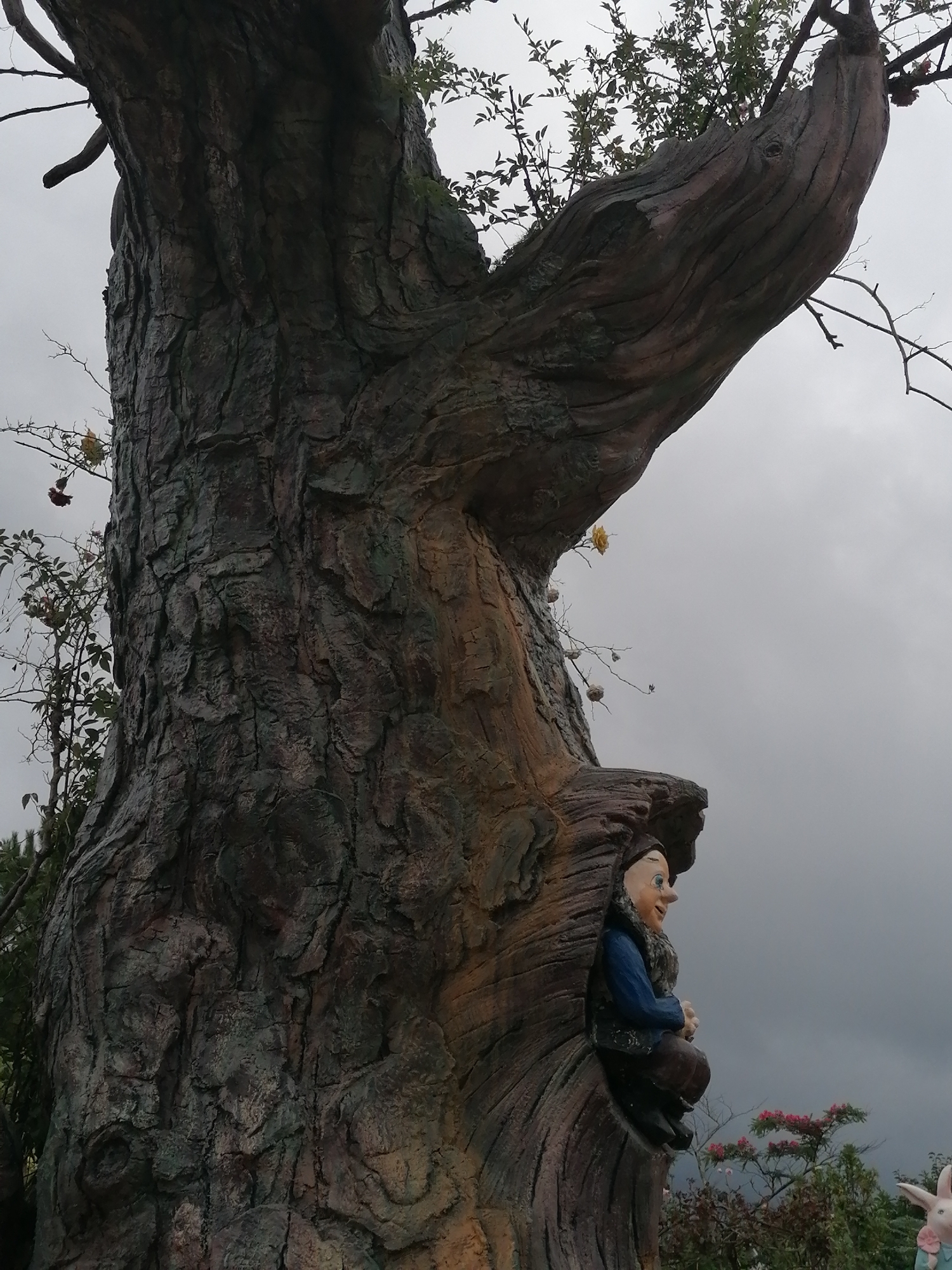 The image captures a whimsical outdoor scene, dominated by a large, artificial tree-like structure with deeply textured, gnarled bark in shades of dark brown and green. The tree's substantial trunk and a thick, upward-curving branch fill much of the frame.
Nestled within a hollowed section of the tree, towards the bottom right, is a figurine resembling a man or gnome. This figure has fair skin, a round blue nose, and a pleasant, smiling expression. He is dressed in a blue shirt, a fur-lined vest, and brown pants, with his hands clasped in front of him.
Sparse vegetation, including small green leaves and a few scattered yellow and dark red flowers, can be seen on branches emerging from or around the tree structure. The sky is overcast and uniformly grey, suggesting a cloudy day. In the very bottom right corner, partially obscured, is the head and upper body of another light-colored figurine, which appears to be a rabbit wearing a blue outfit. The overall setting, located in Lạc Dương, Vietnam, suggests a themed garden or park designed with fairytale elements. There is no visible text in the image.