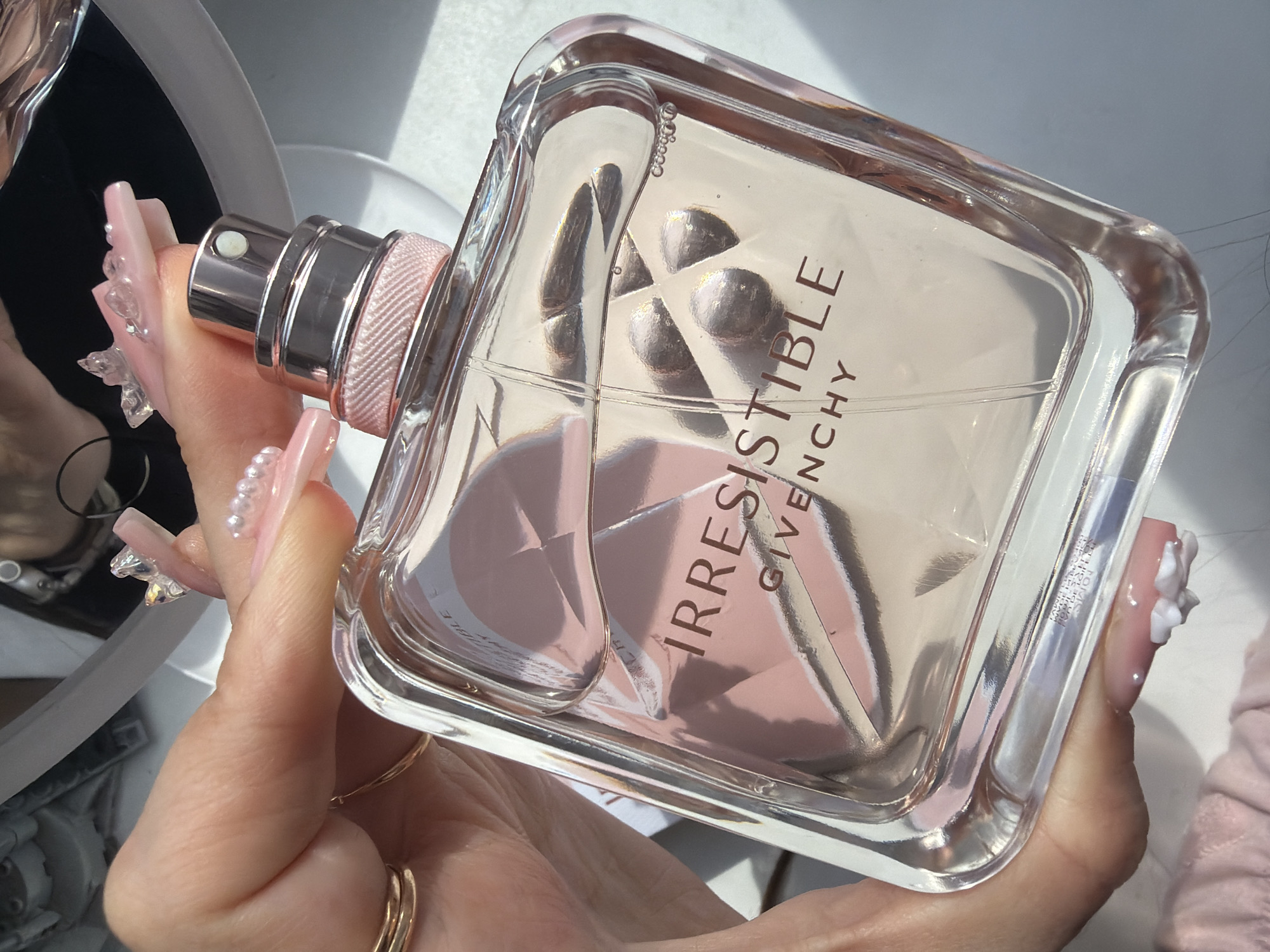 A close-up, eye-level shot captures a person's hand holding a clear glass bottle of "IRRESISTIBLE GIVENCHY" perfume. The bottle is rectangular with rounded corners, containing a clear liquid with a visible fill line and several small air bubbles concentrated near the top right corner. The internal structure of the bottle features a faceted design that creates geometric reflections in shades of soft pink. The metallic silver-toned sprayer is visible at the top, beneath which a textured, light pink ribbon or fabric wraps around the bottle's neck. The product name "IRRESISTIBLE" is prominently displayed in dark reddish-brown capital letters, with "GIVENCHY" in smaller capital letters directly below it, both on the front of the bottle.
The hand has fair skin and is adorned with long, pink-hued, coffin-shaped acrylic nails. The index and ring fingers feature clusters of clear, faceted gems near the cuticles and white, flower-like 3D embellishments near the tips. The middle finger displays a horizontal row of three small, white pearls. Two gold-toned rings, one a simple band and the other with a textured design, are worn on the ring finger. The hand grips the bottle with the thumb on the left side and fingers on the right, partially obscuring the top right portion of the bottle.
The background is softly lit, appearing bright white to the right and bottom of the bottle, suggesting a flat surface like a table or countertop. A distorted reflection, possibly from a mirror or glossy object, is visible in the upper background. The lighting is bright, casting highlights and some soft shadows, indicating an indoor setting likely illuminated by natural light.