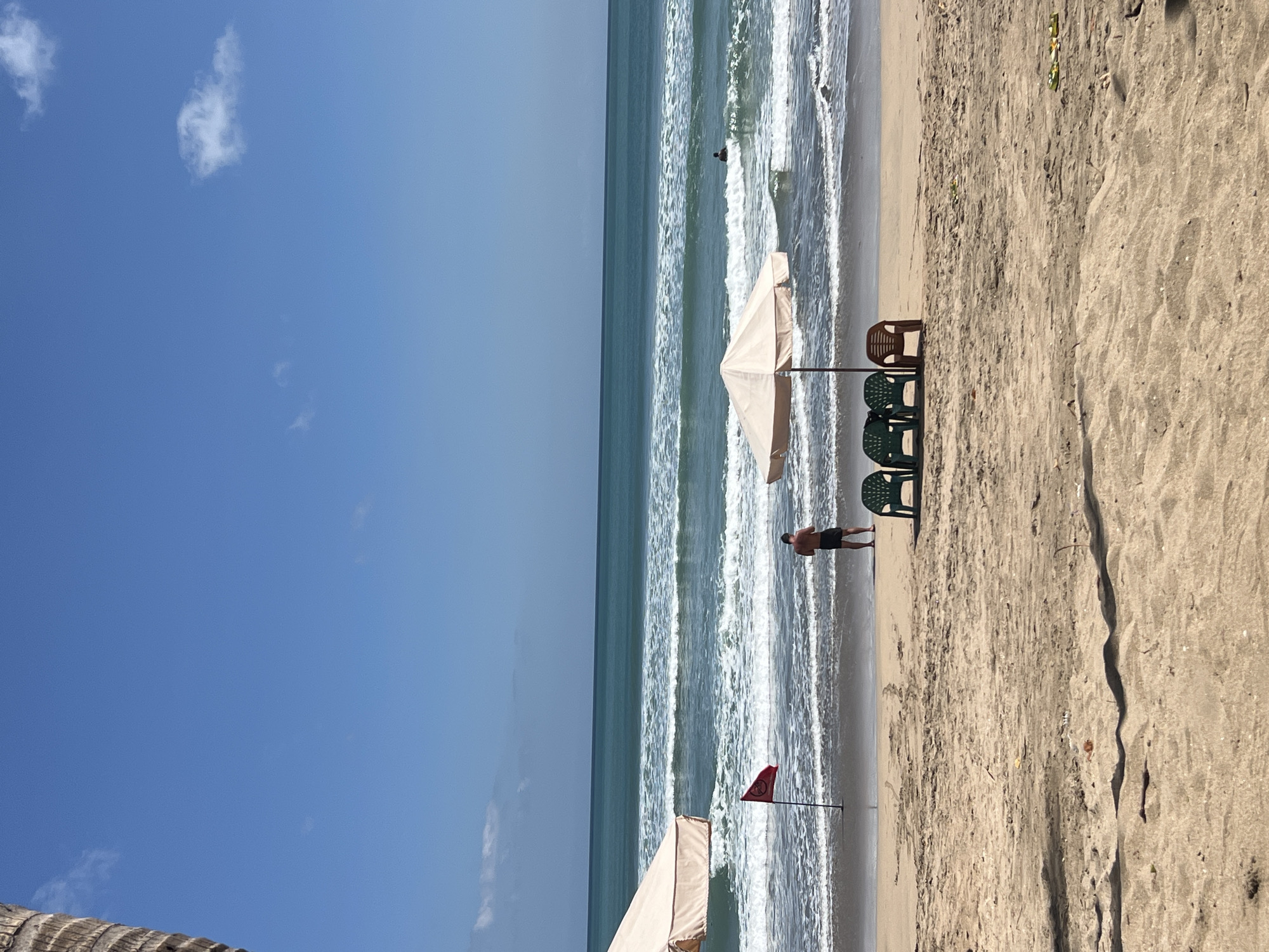 A wide-angle, eye-level, vertical shot captures a serene beach scene in Kuta, Indonesia, under a clear blue sky with sparse white clouds. In the foreground, golden sand stretches across the lower half of the frame, showing subtle indentations and casting long shadows. A row of four plastic chairs, three green and one brown, are neatly arranged under a large, light-colored umbrella, its pole planted firmly in the sand. To the left, partially visible, is another similar umbrella.
The middle ground features the wet sand where the waves gently recede, revealing a red flag planted on a thin pole to the left of center. A solitary man, wearing black shorts, stands on the wet sand facing the ocean, his back to the camera. Further out in the turquoise ocean, waves crash with white foam, indicating a moderate swell. In the distance, a lone swimmer is visible in the water. The horizon line is sharp, dividing the vast expanse of the ocean from the clear sky. A partial view of a textured palm tree trunk is visible on the far left edge of the frame, adding a tropical element to the composition. The overall atmosphere is tranquil, suggesting a typical day at a popular beach resort.