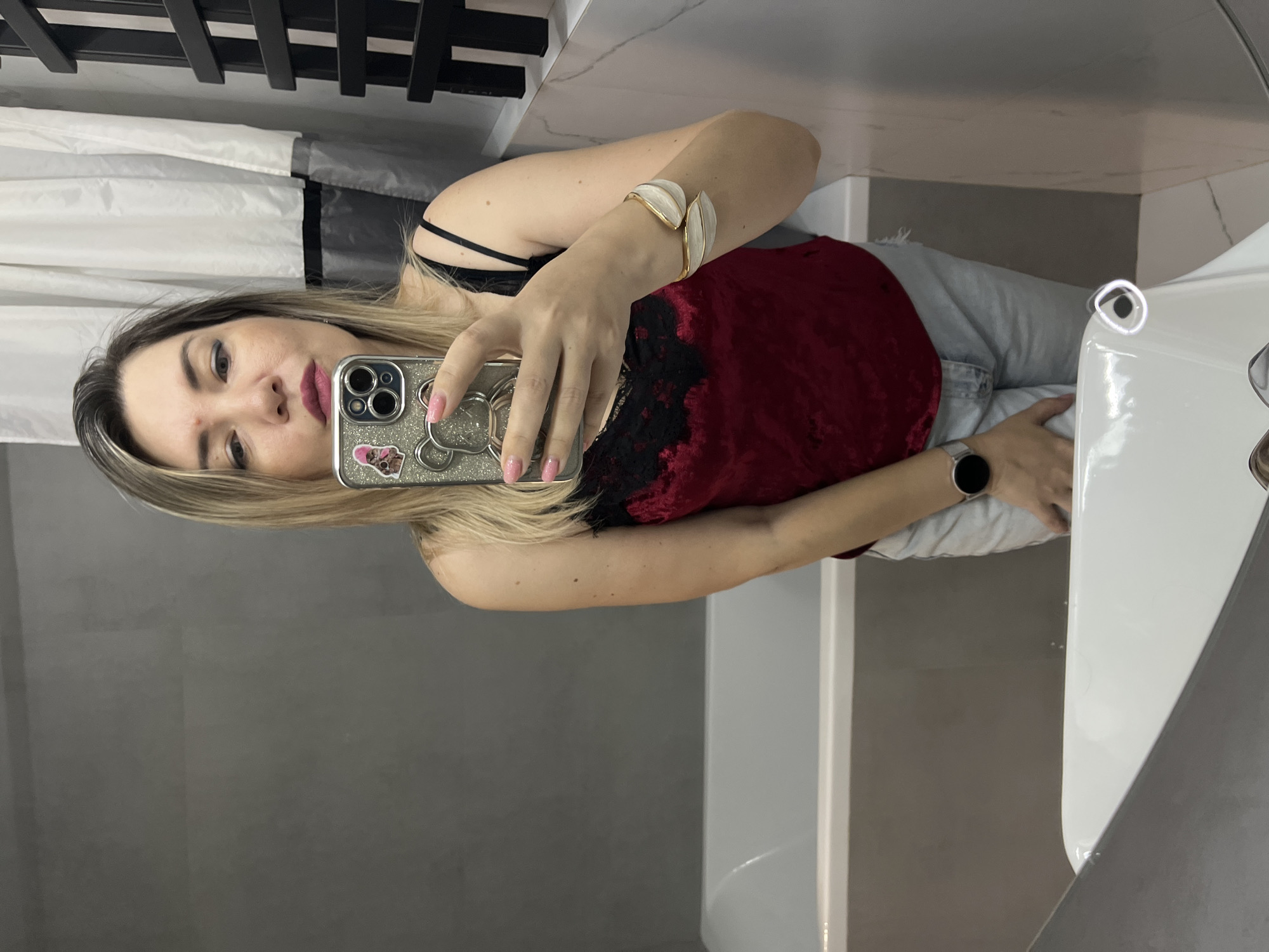 A woman with blonde hair is taking a mirror selfie in a bathroom. She is wearing a red camisole with black lace trim and light-wash denim shorts. She has on a silver-toned bracelet with white leaf-shaped accents and a smartwatch with a black face and silver band. The bathroom features a gray tiled wall, a white bathtub, and a white sink. A white and gray shower curtain is visible behind her. A black radiator-style towel rack is mounted on the wall to the right. The scene appears to be indoors, possibly in a residential bathroom, and there is no direct information in the image to suggest a specific location within the city of Sochi, Russia.