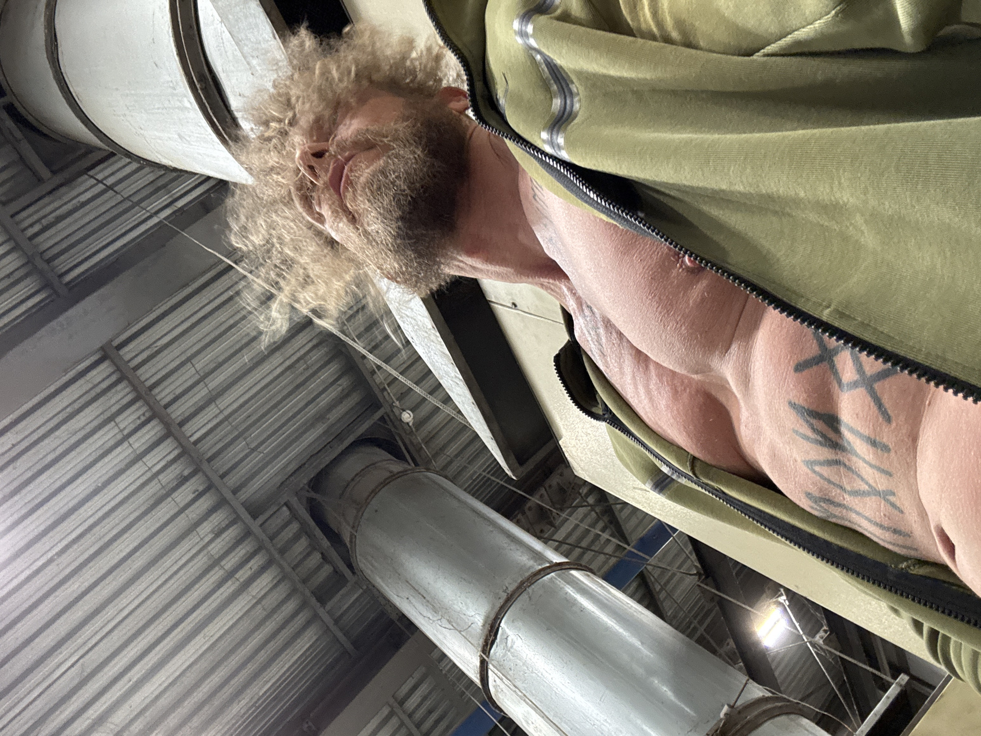 This media file captures a low-angle, upward-looking view of a man within an industrial or warehouse setting in Volzhsky, Russia. The primary subject is a man with light brown, curly hair and a prominent full beard, gazing upwards and slightly to his right. He exhibits a muscular physique, with his bare chest and right arm partially revealed by an unzipped olive green jacket. A distinctive tattoo, resembling stylized angular letters or symbols such as "XXX," is visible on his right side, across his ribcage.
The scene's background features the structural elements of an industrial building. A corrugated metal ceiling with visible support beams spans the upper area. Large, silver-colored cylindrical pipes or ducts are prominent features; one extends vertically from the bottom-center towards the top, while another wider pipe curves into the frame from the top-left. Artificial light fixtures are visible, illuminating the interior. The man's expression appears neutral or contemplative as he observes the overhead industrial architecture. The composition emphasizes the scale and ruggedness of the industrial environment.