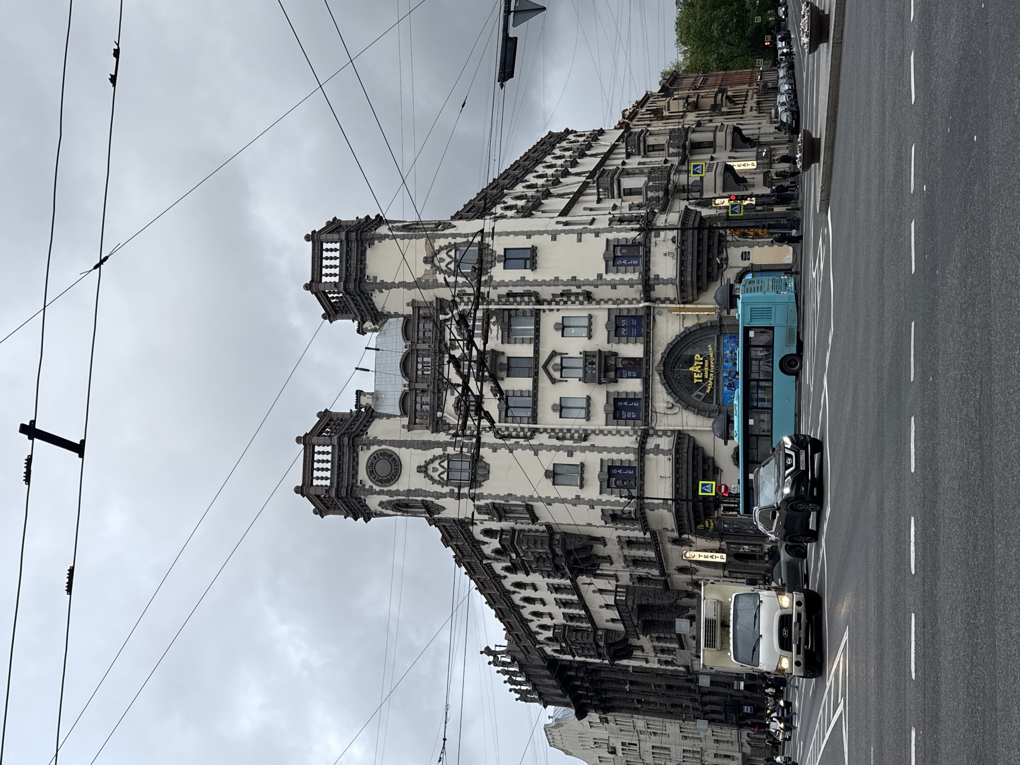 This photograph depicts a street scene in Saint Petersburg, Russia, featuring a multi-story building with elaborate architectural details, including ornate turrets and windows. The building's facade is predominantly white with grey trim. Overhead electrical wires intersect the view of the building. On the street, there are cars including a white box truck, a blue bus, and a dark-colored SUV. There are also road markings for lanes and crosswalks. Additionally, there are traffic signs and signs with Russian script visible on the building, including the word "TEATP" and the Russian word for theatre. The sky is overcast with grey clouds, suggesting a day with potentially inclement weather.