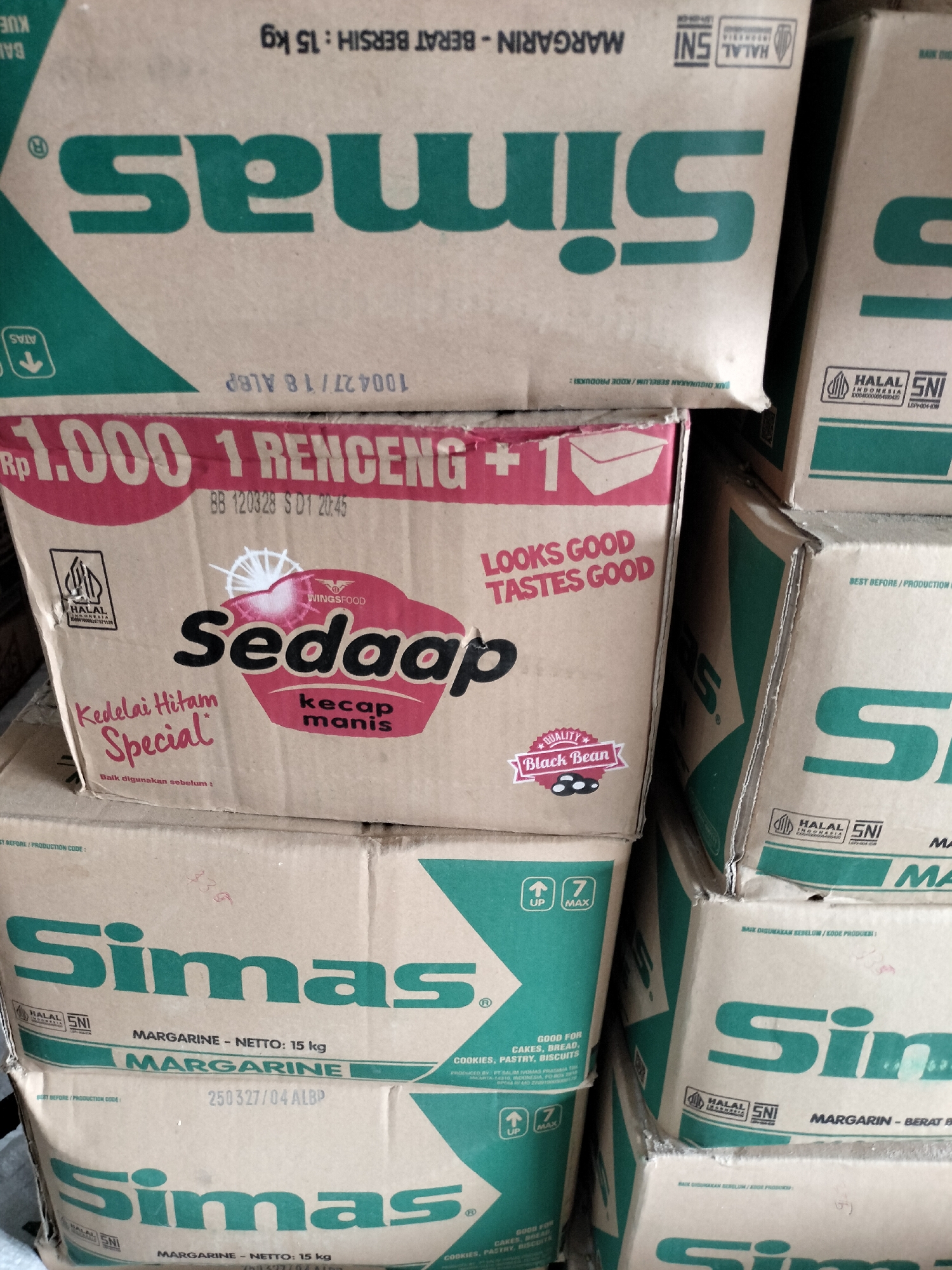 The image shows a stack of cardboard boxes, likely in a storage or retail environment in Meunasahteungoh, Indonesia. The boxes are predominantly white with green and red branding. Several boxes display the brand name "Simas" in large green lettering. Other visible text includes "Margarin - Netto: 15 kg," and product codes. One box features a red and white design with the text "Rp. 1.000 1 Renceng +" and the brand "Sedaap." Another box has the text "Looks Good Tastes Good" and a sticker with "Quality Black Bean." The boxes appear to be manufactured by "Wingsfood." The overall impression is of a commercial inventory.