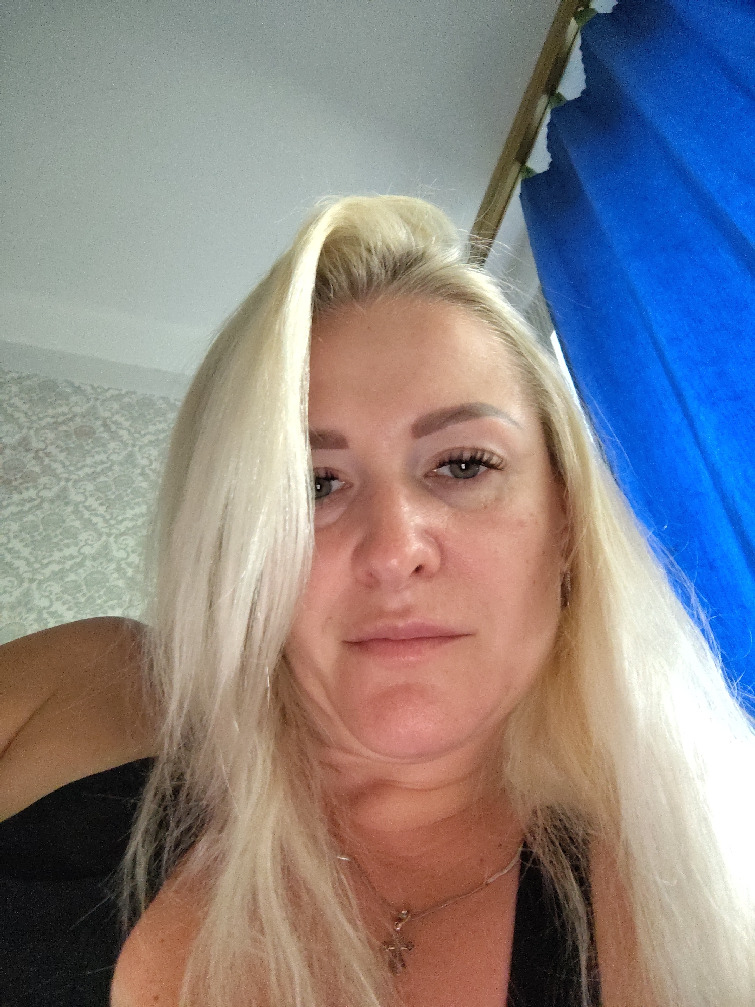 A close-up, selfie-style photograph shows a blonde woman with fair skin and light-colored eyes. She is wearing a black top and a silver necklace with a cross pendant. Her hair is long and straight, framing her face. The background includes a textured, light-colored wallpaper on the left and a bright blue curtain on the right. The lighting suggests an indoor setting. The image appears to be taken in Kyiv, Ukraine.