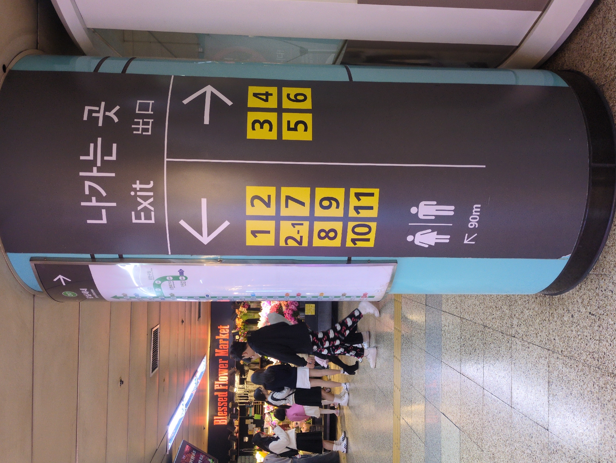 The image captures a busy underground scene in Seoul, South Korea, marked by directional signage and commercial activity. Overhead signage indicates exits 1, 2, 2-1, 3, 4, 5, 6, 7, 8, 9, 10, and 11 with arrows pointing in opposite directions. A sign also points to restrooms 90 meters away. Below the signage is a commercial space with an illuminated sign reading "Blessed Flower Market." A group of students are milling about in the area between the signage and the flower market. The image was taken at 15:36, and the time displayed in the lower left corner is 05:55. Further text on the sign indicates directions to Jamsilsaenai, Sinjeongnegeori, Kkachisan, and Yangcheongu Office.