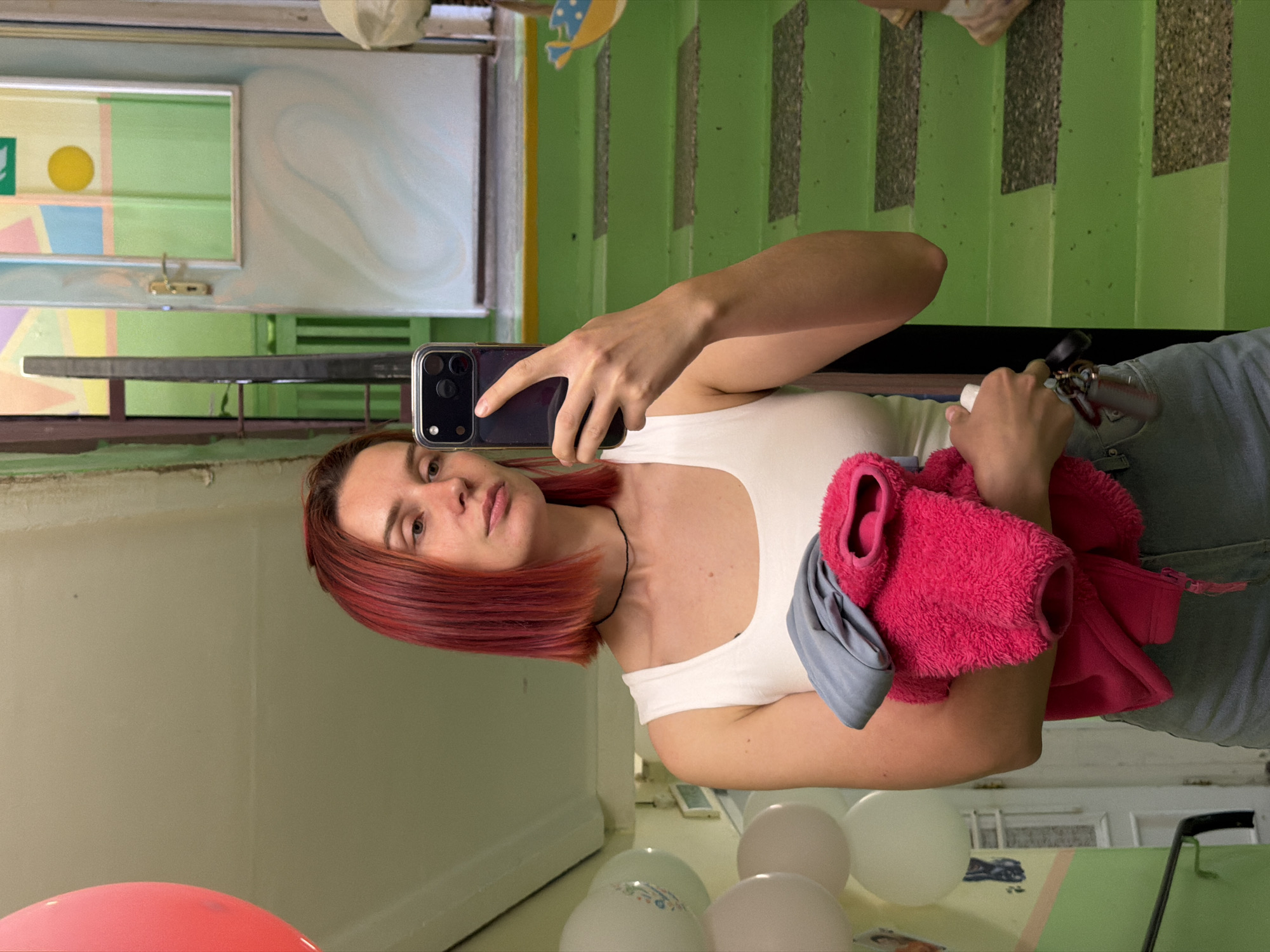 A red-haired woman takes a selfie in what appears to be a stairwell or hallway. She is holding a smartphone in her right hand and a pink fleece jacket in her left. The environment features green-painted stairs, a mirror, a pink balloon, and several white balloons on the floor. The location is in the city of Chaikovsky, Russia.