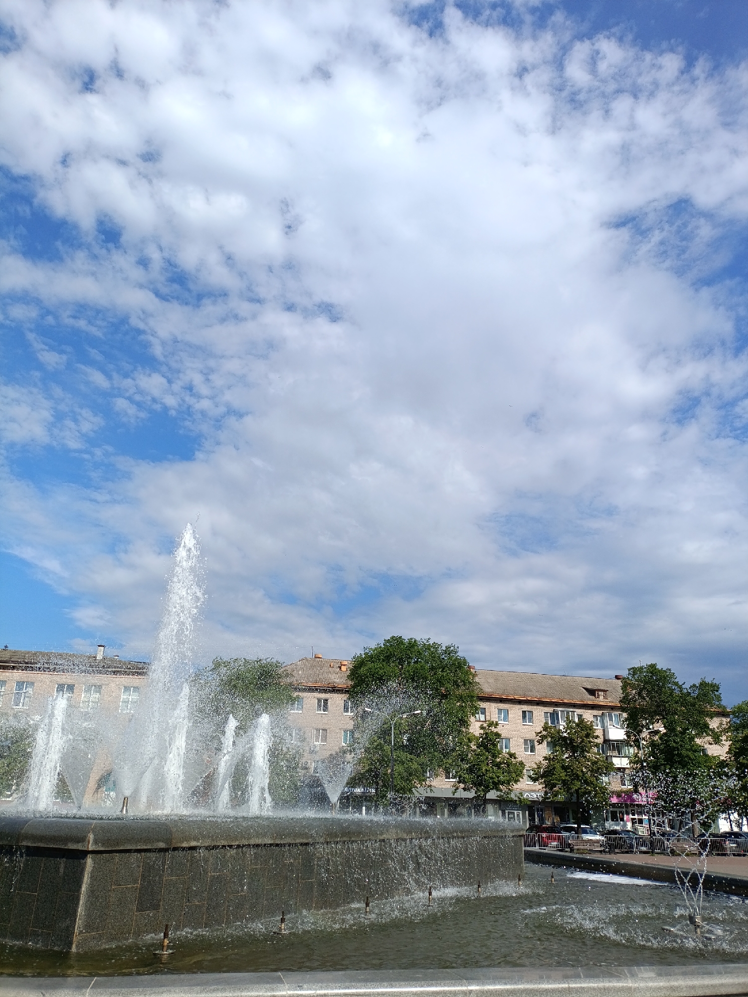 The image depicts a fountain in the city of Chaikovsky, Russia. The fountain features multiple jets of water spraying upwards, set against a backdrop of a multi-story building with a grey tiled roof. Trees with green foliage are interspersed between the building and the fountain. The sky above is blue with numerous white clouds. Cars and pedestrians are visible on the street behind the fountain. The fountain is made of dark gray stone.