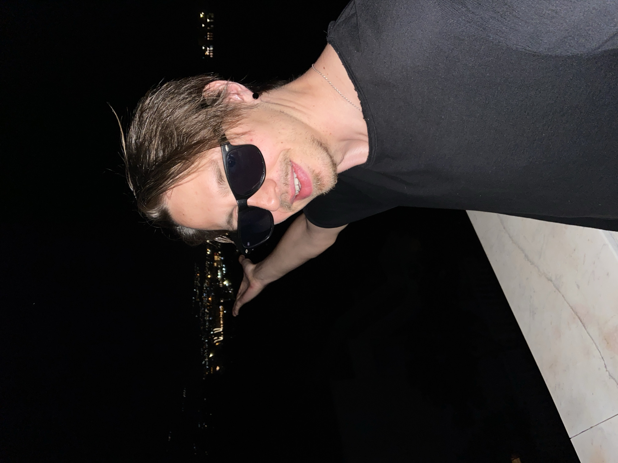 A man with brown hair and sunglasses is positioned in the top half of the image, leaning toward the camera. He is wearing a black t-shirt and a silver chain. His arm extends towards the bottom left of the image, with the city lights of Šušanj, Montenegro, blurred in the background. The setting is at night. In the bottom right corner, there is a white marble or tiled surface.