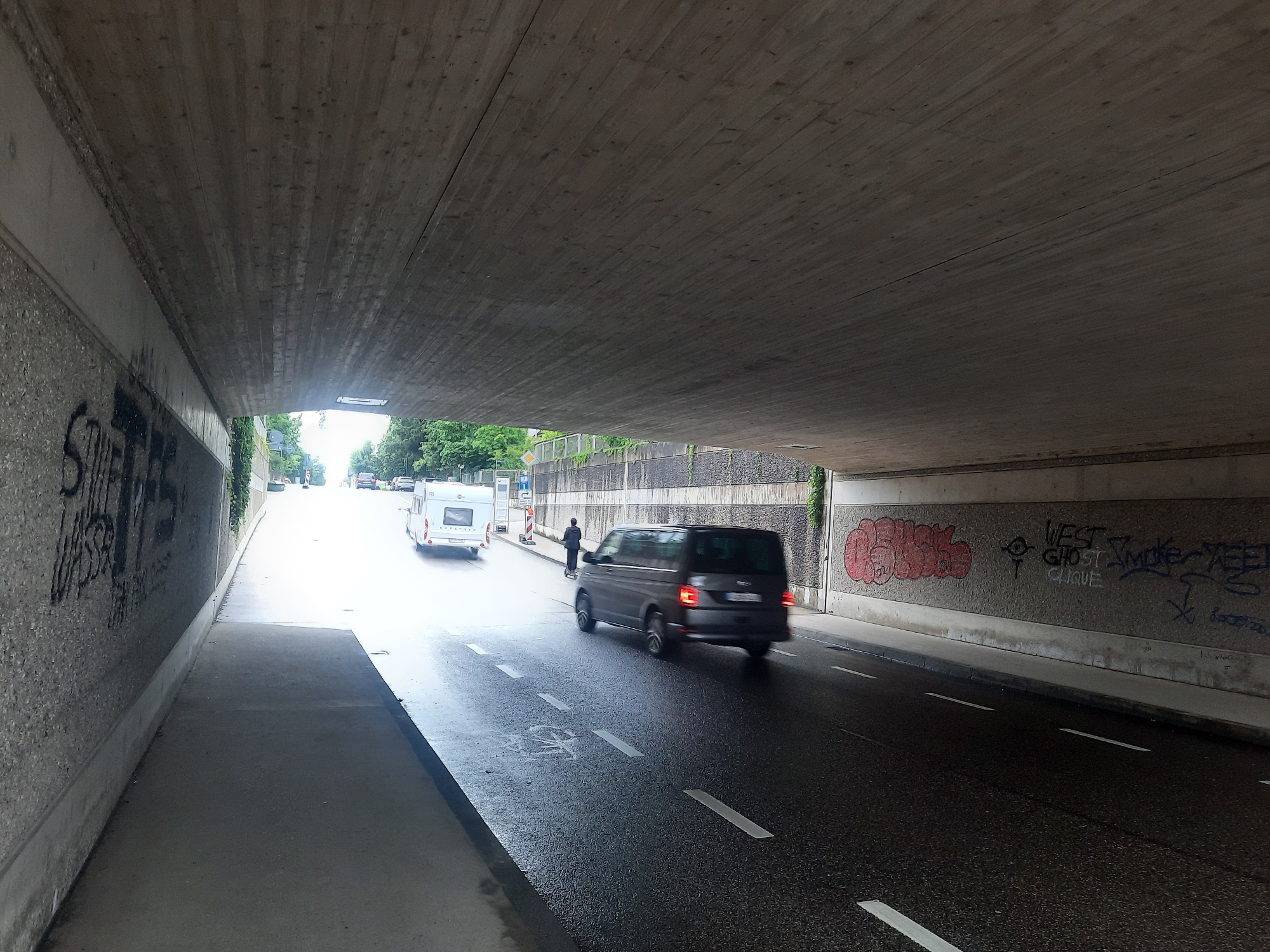 A dark, concrete underpass is visible, with graffiti on the left wall. A paved road runs through the underpass, with a pedestrian walkway on the left. A dark gray van is moving away from the camera on the right side of the road. On the left side of the road, a white caravan is also moving away from the camera. A person is visible on a scooter in front of the van. The ceiling of the underpass is made of concrete with visible wooden formwork impressions. Sunlight is entering the underpass from the far end, creating a bright light. The walls of the underpass are lined with concrete and show various graffiti tags, including red spray-painted text on the right wall. The road surface appears wet.