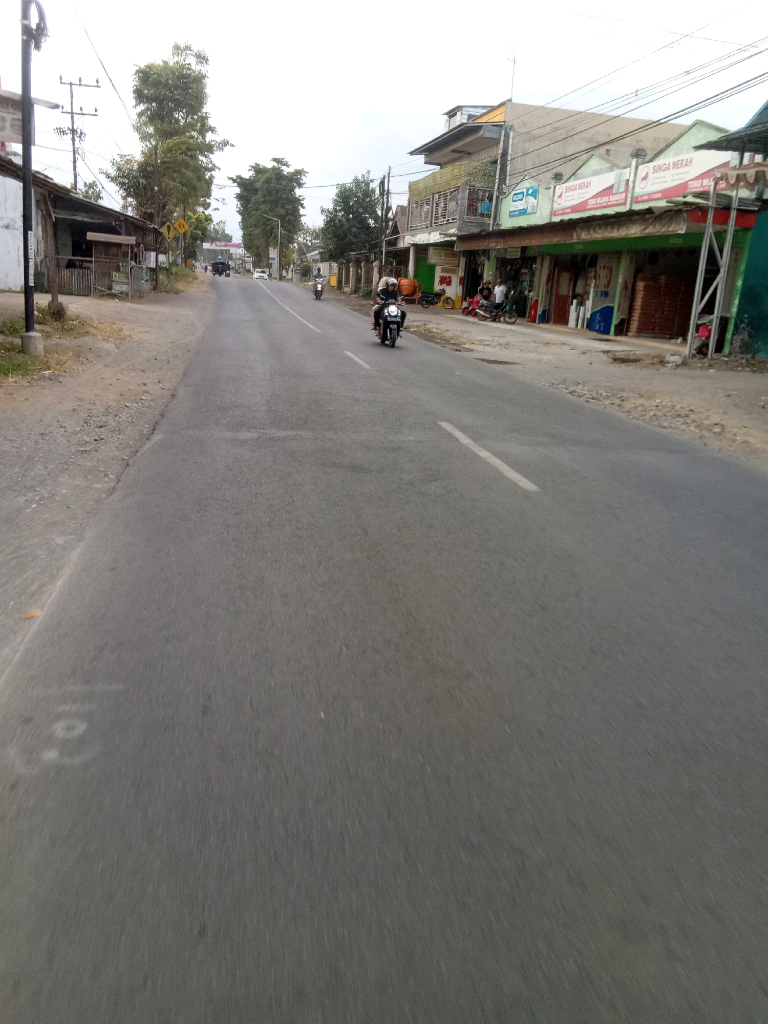 The image depicts a street scene in Ajung, Indonesia, likely in a semi-urban or rural area given the presence of shops and a paved road. The perspective is from a moving vehicle, giving a sense of forward motion. On the left side of the road, there are simple structures, possibly shops or houses, with one appearing to be a small, weathered building with a corrugated metal roof. A utility pole with wires is visible. Further down the road, there are more substantial buildings, some with signage in what appears to be Indonesian, indicating businesses. One building prominently displays the name "SINGA MERAH." Several motorcycles are present; one is being ridden by a person in the foreground, and others are parked or being ridden further down the road. A few individuals are seen near the shops. The road itself is paved with a white line marking the center. The environment appears to be a commercial strip with a mix of residential and business establishments lining the street, with vegetation and trees interspersed. The sky is overcast.