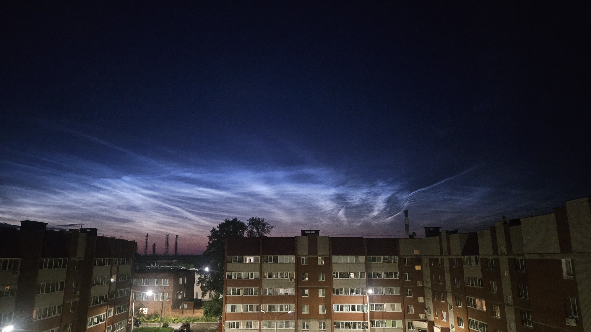 The image is a nighttime, wide-angle view of an urban landscape under a dark blue sky filled with noctilucent clouds. The clouds are wispy and luminous, illuminated from below by the last vestiges of sunlight or artificial light. In the foreground, the tops of several multi-story apartment buildings are visible. These buildings are constructed with reddish-brown brick and have numerous windows, some of which are lit. Streetlights are visible at ground level near the buildings, casting a yellow glow. In the middle ground, silhouetted trees stand between the buildings. Further in the background, several tall industrial towers with lattice structures are visible on the horizon, suggesting an industrial area on the outskirts of the city. The overall impression is one of a quiet, somewhat desolate, but visually striking urban scene at dusk or early night, highlighting the natural phenomenon of noctilucent clouds. The location is identified as the city of Glazov, Russia.