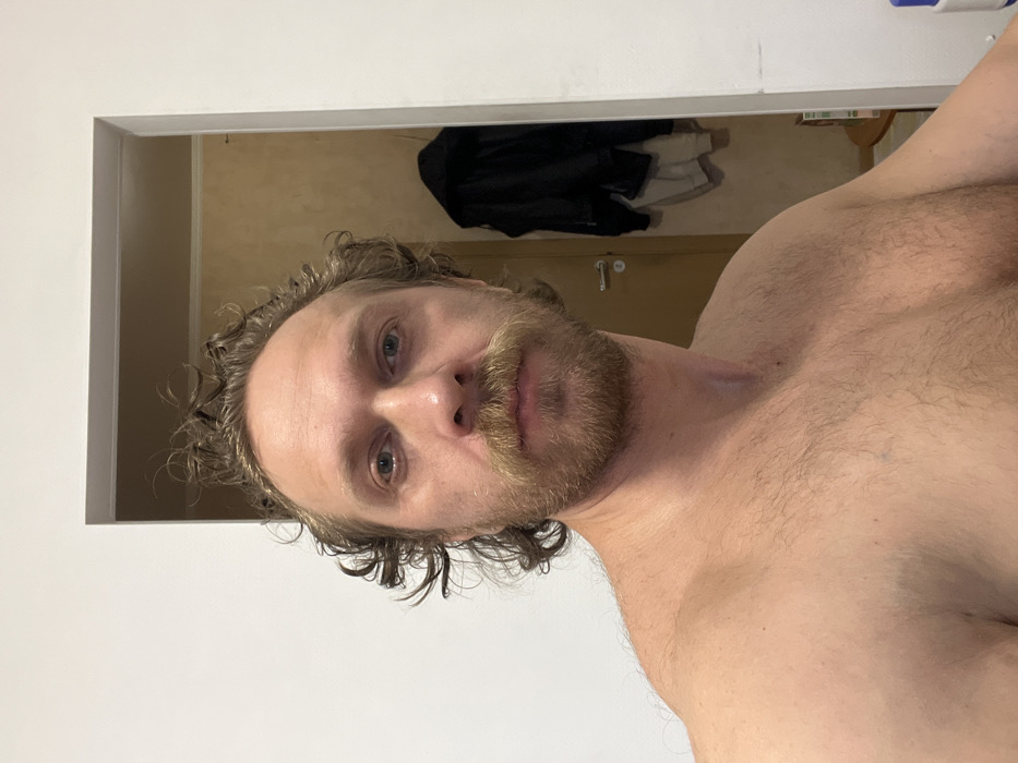 A shirtless man with light skin, a beard, and curly hair is facing the camera. He stands within a doorway against a light beige wall. Behind him, visible through the doorway, hang a black jacket and a white garment. A metallic door handle is on the wall behind him, and there's also a hint of a chair in the background.