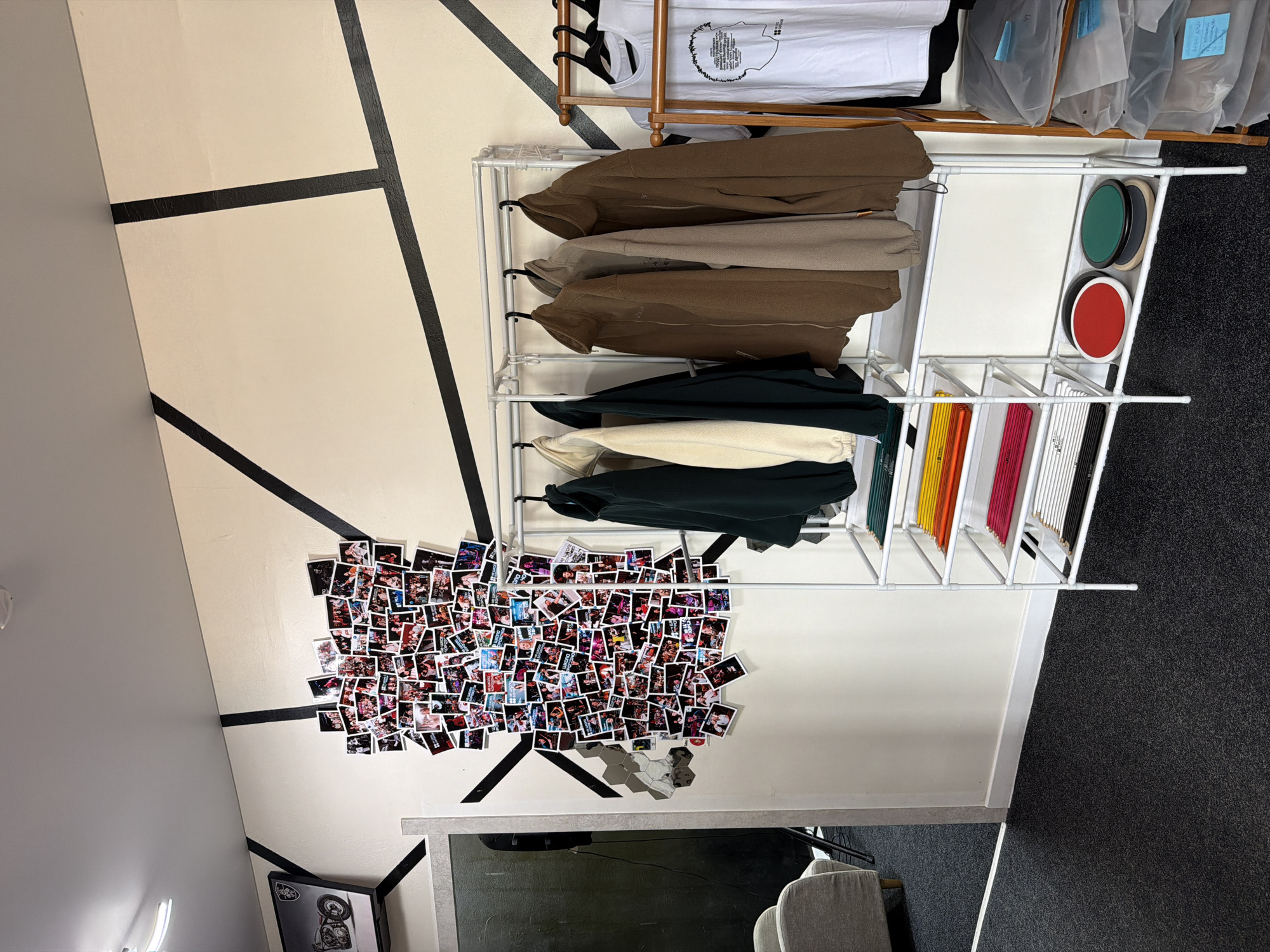The image shows a room with a minimalist aesthetic, featuring a wall with a cream background and bold black geometric lines. On the wall, a collage of small photographs is displayed, depicting a variety of people and scenes, possibly from personal memories or events.
Adjacent to the wall is a white metal shelving unit. The top shelf holds several white t-shirts, some appearing to be packaged. Below this, multiple rows of clothing are neatly hung on hangers, including several hoodies in earthy tones like brown and beige, and some darker colored garments. The lower shelves of the unit contain various items, including a stack of colorful folders or binders (yellow, orange, pink, and black) and some circular objects (one green, one beige, and one red) stacked in a compartment.
The room appears to be an indoor space, possibly a bedroom, office, or studio, given the presence of the shelving unit and the decor. The lighting suggests it is daytime, but no specific weather conditions are discernible. The overall mood conveyed is one of organization and personal expression, with the photo collage adding a touch of personality to the otherwise clean and structured environment. There is no discernible text in the image that provides specific information about the location or context beyond what can be inferred from the visual elements. The provided location context of Petropavlovsk-Kamchatsky, Russia, does not yield any further specific visual cues within the image itself.