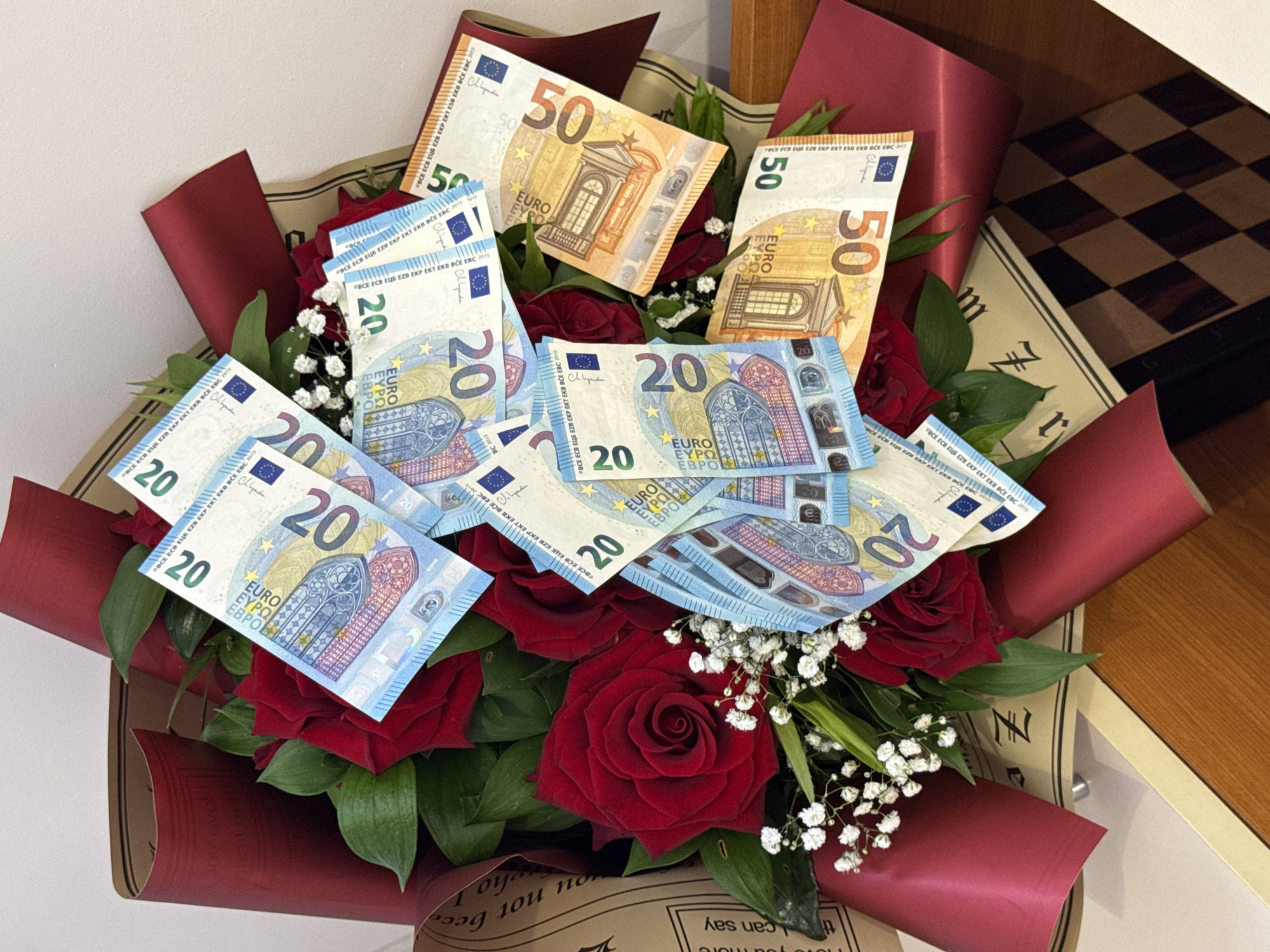 This close-up, high-angle shot features a vibrant bouquet presented indoors. The central subjects are numerous deep red roses, complemented by delicate white baby's breath flowers and lush green foliage. The bouquet is stylishly wrapped in a combination of dark red and patterned brown paper, adding to its festive appearance.
Uniquely, the bouquet is interspersed with Euro banknotes, acting as decorative elements. Several blue-hued 20 Euro notes and golden-orange 50 Euro notes are prominently displayed among the flowers. The notes show security features and standard text like "EURO," "EYPΩ," "EBPO," and "BCE ECB EZB EZB EXP EKT EKB BCE EBC 2015," along with a visible signature.
In the background, a corner of a wooden chessboard with dark and light squares is partially visible to the right, suggesting a recreational or domestic setting. A plain white wall forms the upper left background. Text is also visible on the brown wrapping paper, with fragmented English phrases such as "you no," "ot beco," "who," and "can say," along with other illegible characters, hinting at a decorative or meaningful message. The arrangement conveys a sense of celebration or a special gift, blending traditional floral beauty with a practical monetary contribution.