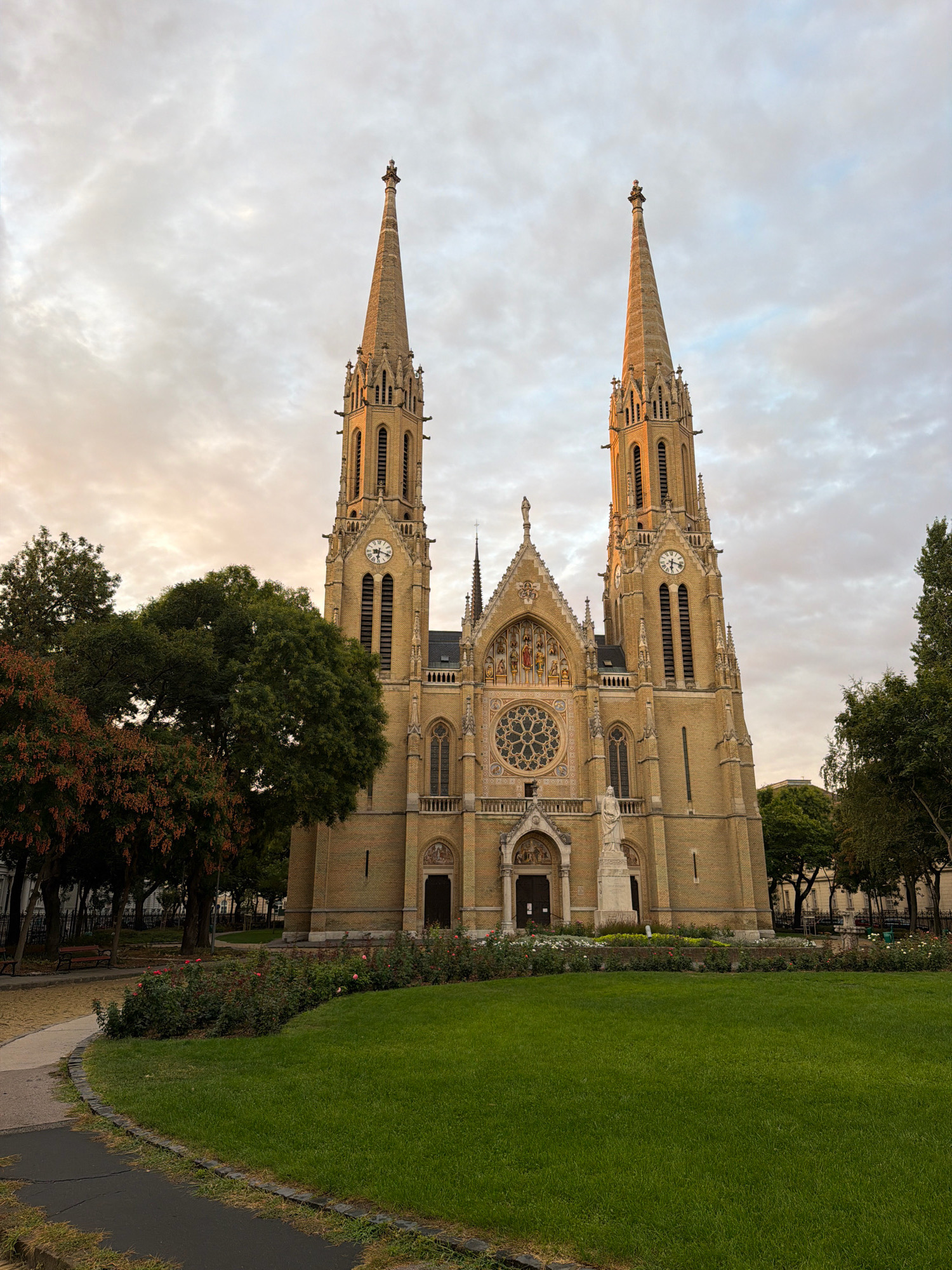 The image shows the exterior of St. Elizabeth of Hungary Church in Budapest, Hungary. The church is a large, yellow brick building with two prominent spires, a clock on each tower, and a rose window above the entrance. In front of the church is a well-maintained lawn and a flowerbed with roses. Trees surround the church and the park area. The sky is cloudy, and the light suggests it is either early morning or late evening. There are no people visible in the image.