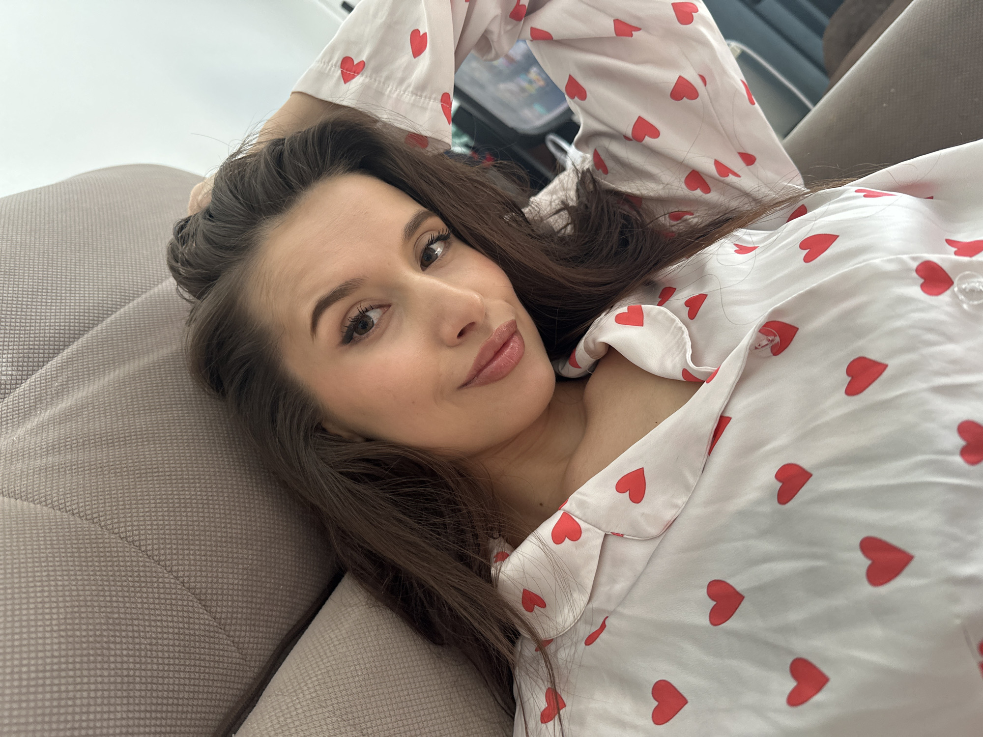 A woman is reclining on a beige couch, wearing a white button-down pajama top adorned with red heart patterns. Her dark brown hair is partially tucked behind her head, and she is looking directly at the camera with a slight smile. The setting appears to be indoors, likely a private residence. There's no specific information in the image to indicate the location is Yekaterinburg, Russia.
