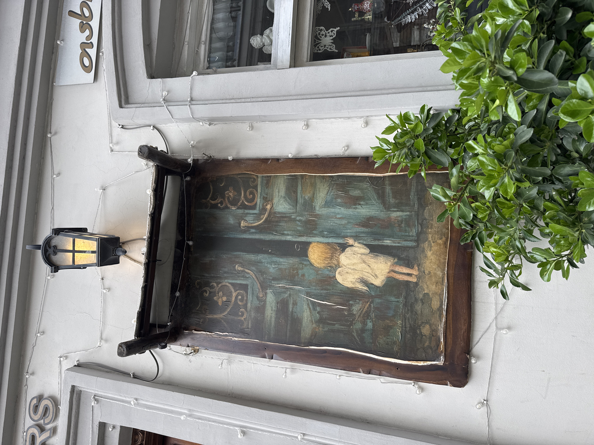 A view of an outdoor wall of a building features a framed painting of a child walking towards an ornate, teal door. The wooden frame of the painting is supported by two black, wooden brackets. To the left is a black metal lantern affixed to the wall. A bush with glossy green leaves grows to the right of the painting, and a string of small, white lights are visible around the perimeter of the frame and on the wall. Above the painting is a window with a white frame. A partial sign with the letters "ons" in gold can be seen on the upper left corner of the wall. The location is Unknown, Unknown.