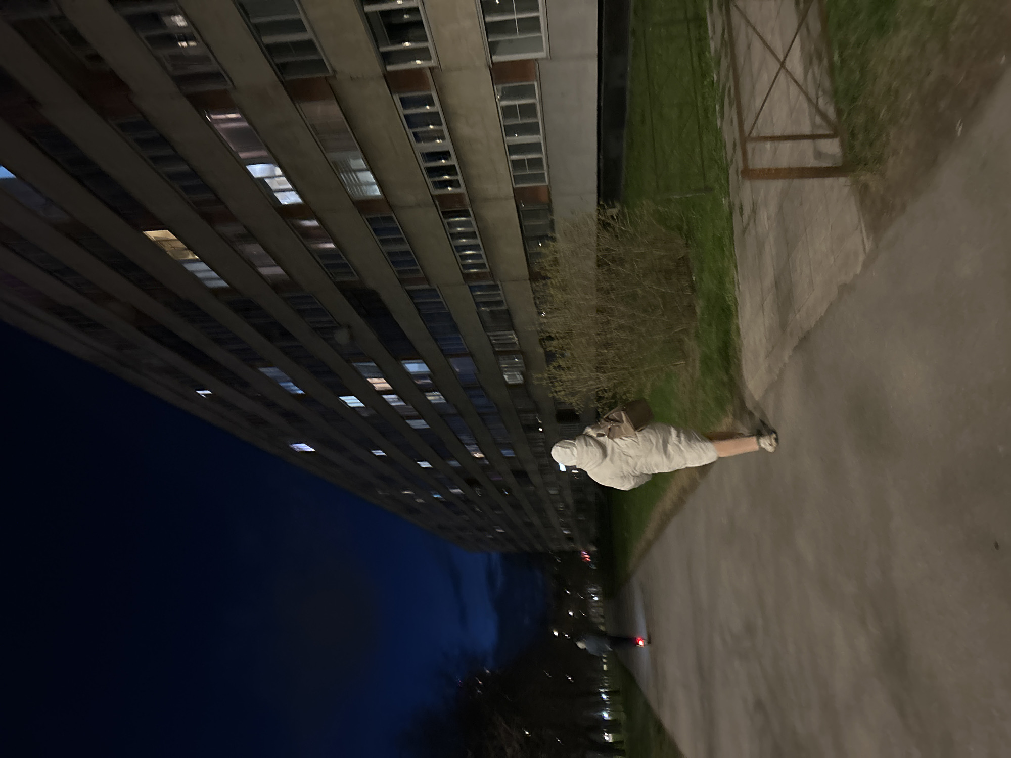 This image captures an urban street scene in Saint Petersburg, Russia, at dusk or night. The photo is taken in portrait orientation but depicts a vertical building on the left and a horizontal ground plane on the right.
Dominating the left side is a tall, multi-story building, likely residential or office, characterized by a grid of numerous windows set into a light-colored facade. Many of the windows are illuminated, casting light and reflections across the dark building surface. The architectural style appears functional, reminiscent of Soviet-era or post-Soviet structures.
In the mid-ground and foreground, a paved pathway or sidewalk stretches horizontally, appearing to be concrete or asphalt. A strip of grassy land with sparse, bare bushes and trees runs parallel to the path. A rustic metal fence or railing, with an X-pattern, is visible along the edge of the grass closer to the building.
A lone person stands on the paved path in the foreground, wearing a light-colored, possibly white or light grey, hooded garment that obscures most of their features. A bag or backpack is visible slung over their shoulder. The person is oriented away from the viewer, looking down the path. Further down the pathway, in the blurred distance, a small red light suggests another person, possibly on a bicycle, or a distant vehicle.
The sky is a dark blue, transitioning to black, indicating late evening or night. The overall lighting is dim, relying on the artificial lights from the building windows and potentially distant streetlights. The scene conveys a quiet, solitary atmosphere. No visible text is present in the image.