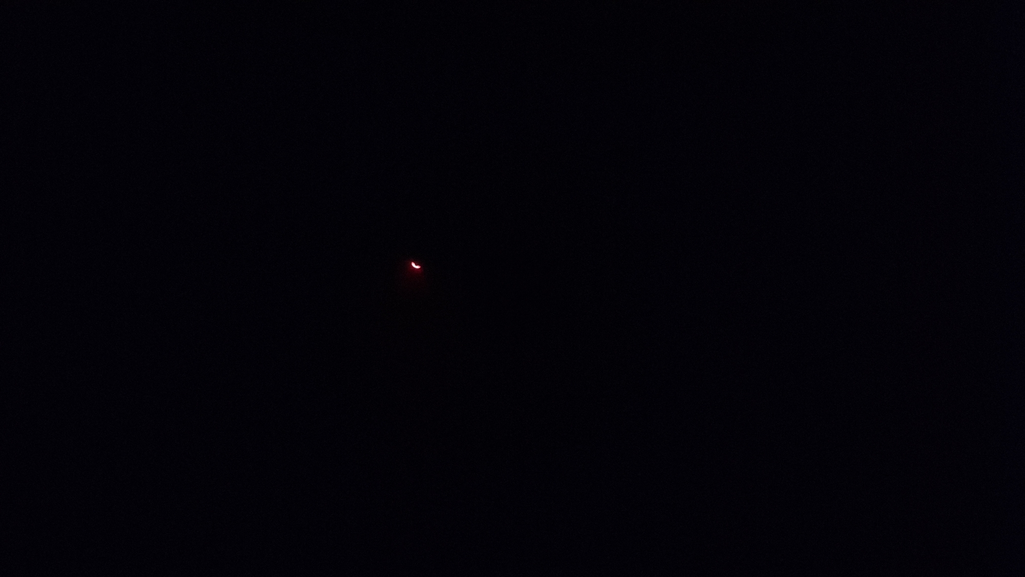 The image shows a partial solar eclipse, with a sliver of the sun illuminated, appearing as a red crescent against a dark sky. The event is taking place in Híjar, Spain. The darkness suggests it is either nighttime or the eclipse is occurring during daytime, obscuring the sun. There are no people, objects, or other scene elements visible, leaving the focus entirely on the celestial phenomenon. No text is present in the image.