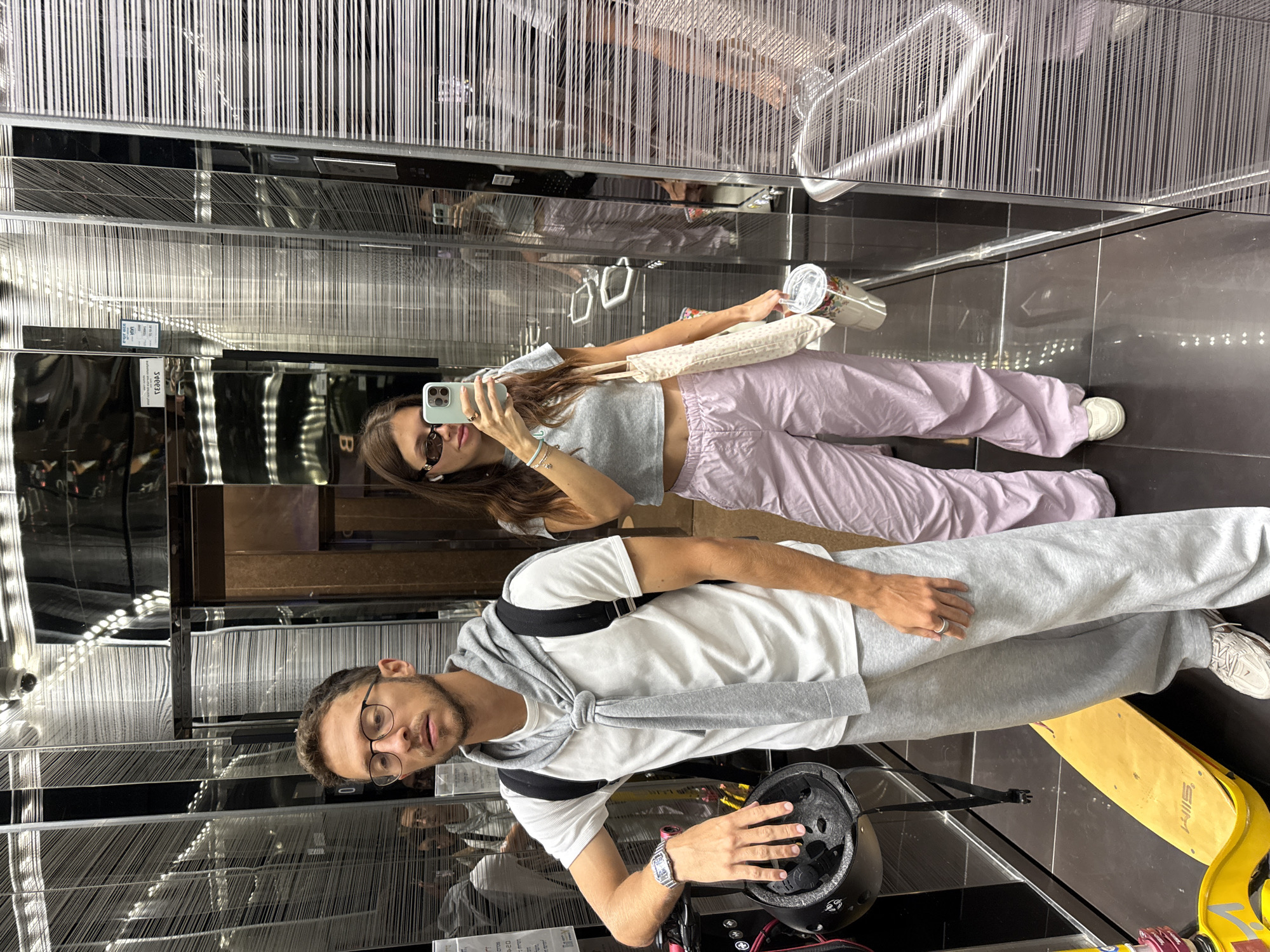In a modern elevator with reflective walls and decorative vertical strands, a man and a woman pose for a selfie. The woman holds a smartphone, wearing sunglasses, a gray crop top, and lilac sweatpants, with a shoulder bag and a drink in her other hand. The man wears glasses, a white t-shirt, gray sweatpants, and a gray sweater tied over his shoulders. He is holding a helmet. A yellow scooter rests on the floor near the man. The location is a modern building in Tel-Aviv, Israel.