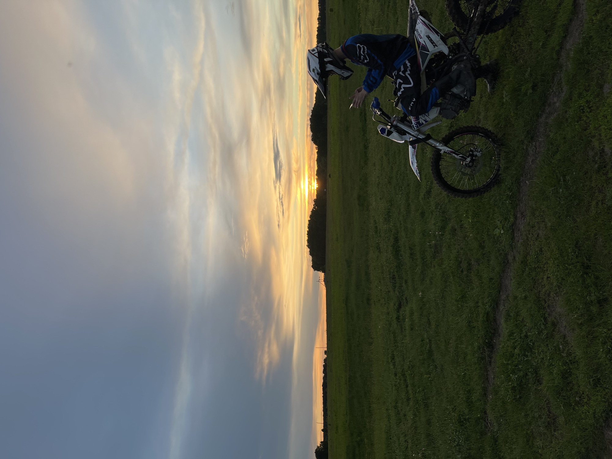 The image shows a person on a motorcycle in a grassy field, with a sunset in the background. The person is wearing a helmet and riding gear. The motorcycle is white with black and blue accents. The grass appears to be in a field with some paths, possibly in the region of Новочувашский, Russia. The sky is partially cloudy, with colors of orange, yellow, and blue indicating a sunset.