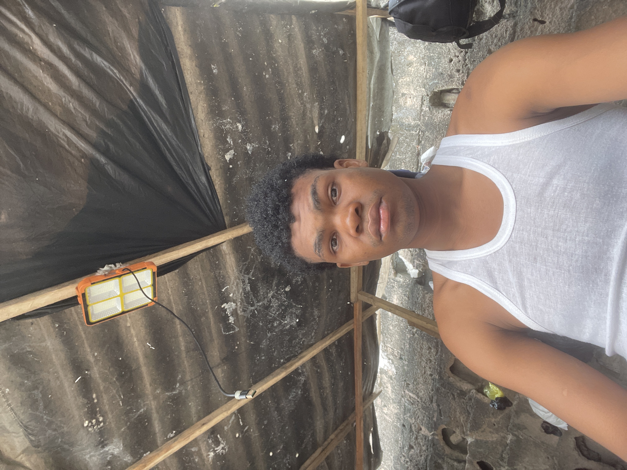 The image is a medium shot selfie of a young Black male wearing a white tank top. He is positioned centrally in the frame, looking directly at the camera with a neutral expression. His hair is dark and curly, styled short. The background appears to be an outdoor, possibly semi-constructed, shelter. Above him, a corrugated roof is visible, covered in parts by black plastic sheeting. A bright LED work light is attached to the roof structure, illuminating the scene. Wooden beams support the roof. To the left and right of the subject, parts of a rough, textured wall are visible. A black backpack is slung over a post on the right side of the frame. The overall environment suggests a utilitarian or temporary setting, possibly in a developing area. The reference to Ilado, Nigeria, provides a specific geographic context for this scene.