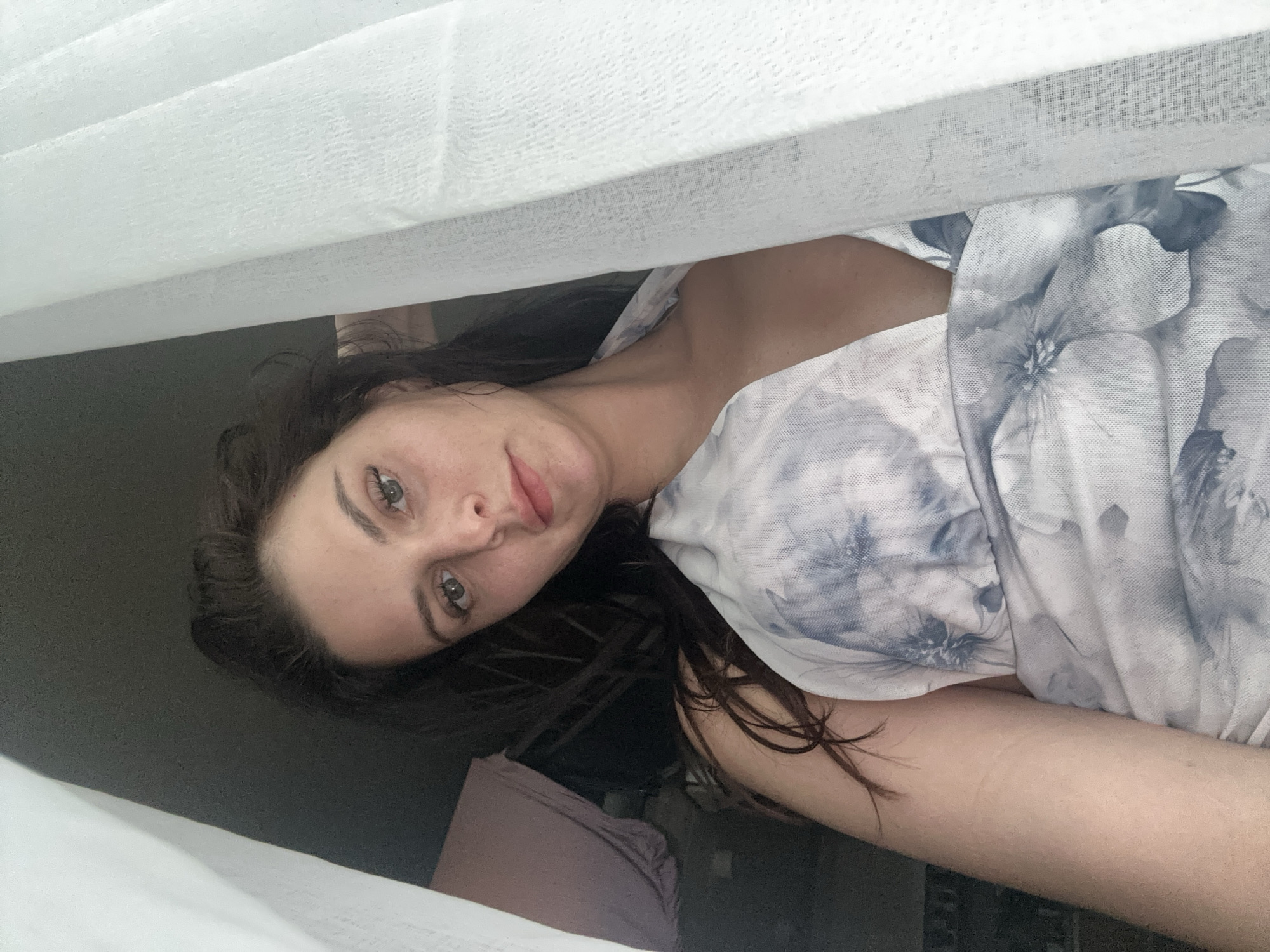 A medium shot of a woman with long, dark hair is captured from a low angle, looking upwards towards the camera. She is positioned behind sheer white curtains, which partially obscure her face and body. Her hair is loosely styled, with some strands falling around her face. She is wearing a light-colored dress with a floral pattern of blue and white. Her eyes are looking directly at the camera, and she has a neutral expression. The background is dark and out of focus, with some indistinct objects visible, suggesting an indoor setting. The lighting is soft and diffused, likely from a window or natural light source.