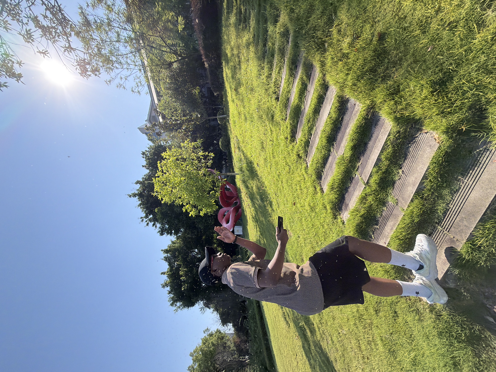In a park in Jiangjin, China, a Black man in a striped polo shirt, black shorts, white socks, and white sneakers stands on a grassy slope next to a set of wooden steps. He wears a black baseball cap and is looking up and to his right. In his right hand, he holds a black remote control, and his left hand is raised as if he is swatting at something. The sun is bright in the sky, and there are many small, white specks that appear to be insects or dust in the air. In the background, there are trees, a building, and a large red art sculpture. The grass is green and well-maintained, and the wooden steps are partly covered by grass. The scene is illuminated by bright sunlight, casting shadows on the grass.