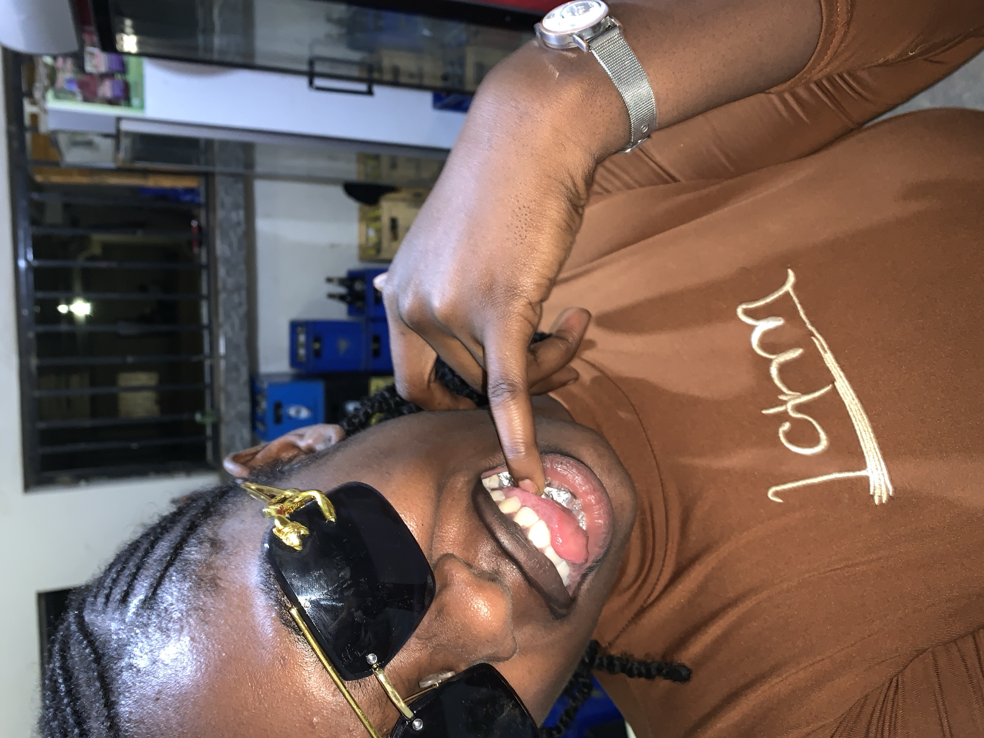 A high-angle, medium-close shot depicts an individual posing in an indoor commercial environment located in New Stadium, Nigeria. The subject, a person with dark skin, wears braided hair and black sunglasses featuring prominent gold, animal-shaped accents on the frames. They are dressed in a brown, high-neck, long-sleeve top with "LCNM" or similar white embroidered lettering on the chest. A silver-toned watch with a mesh strap is visible on their left wrist. The individual's left index finger touches their lower lip, while their tongue is extended over a metallic dental covering or grill on their lower front teeth. The background features elements of a retail or storage space, including stacked blue plastic crates and brown glass bottles on the left. To the right, a tall, white commercial refrigerator with a black handle is visible, alongside another dark-framed vertical structure. Above these, a window or opening secured with dark metal bars reveals a dimly lit area beyond.
