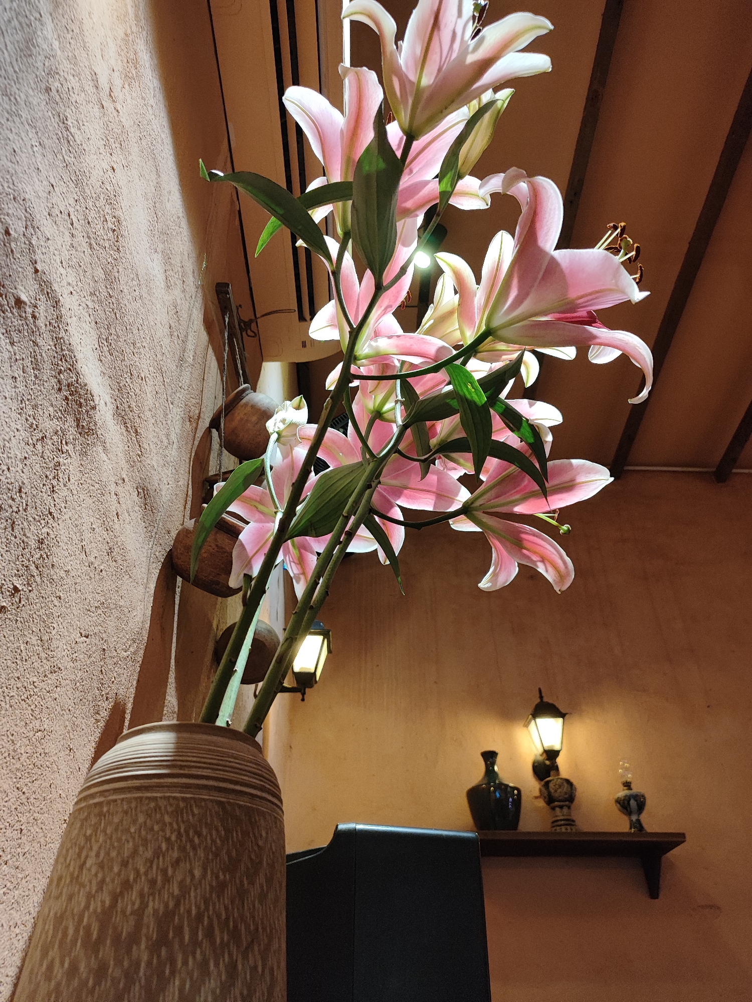 A close-up, low-angle shot shows a cluster of pink lilies in a large, speckled ceramic vase against a textured, terracotta-colored wall. The lilies are the primary subject, with their delicate petals catching the light from above. The background features a sloped, wooden ceiling, a small wall-mounted lamp, and a shelf adorned with decorative vases and another lamp. The overall impression is one of warm, ambient lighting and natural beauty. The setting is located in Da Nang, Vietnam, likely a restaurant or home, given the decorative elements and the style of the room. No people are present in the image.