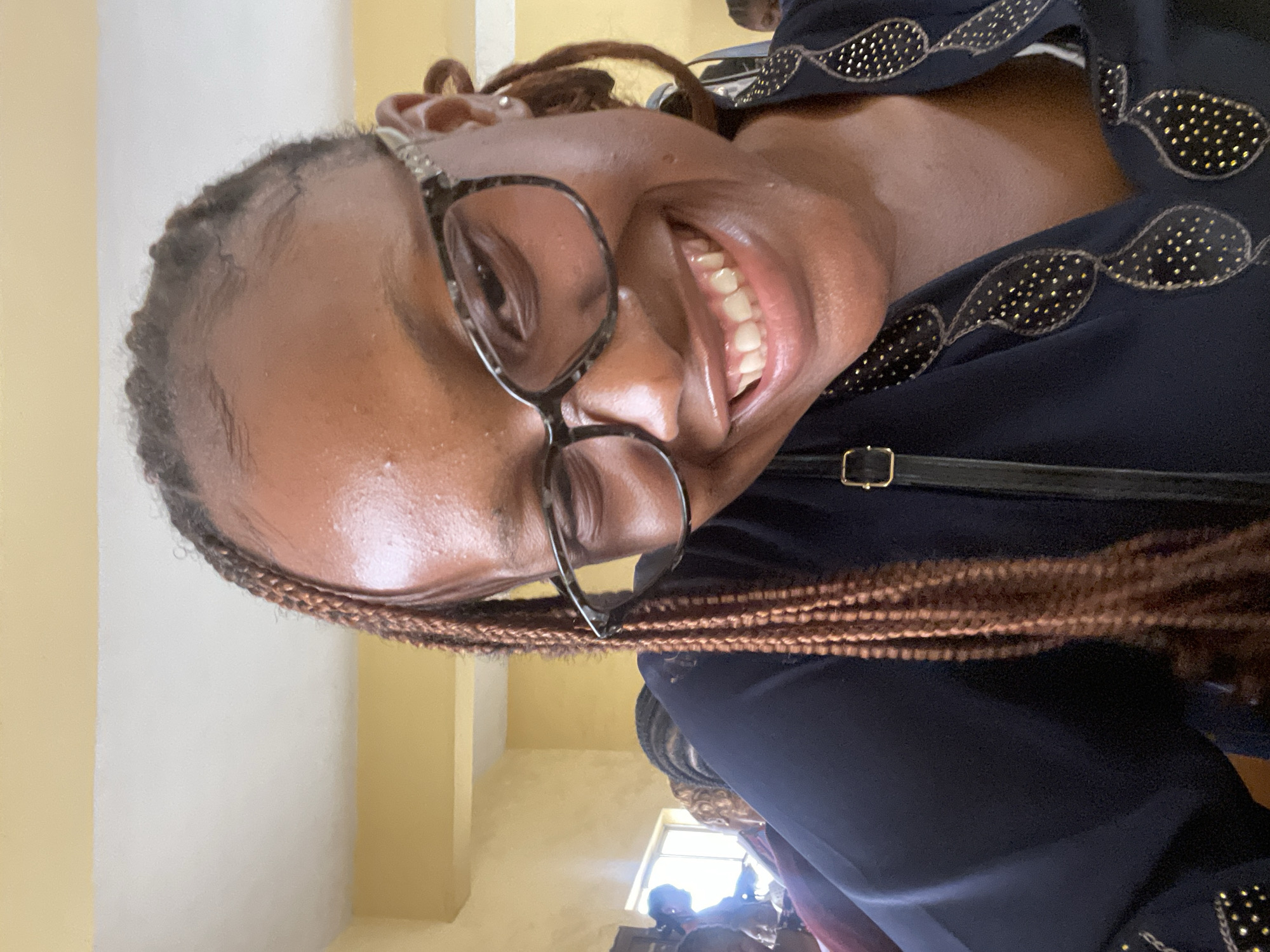 The media file is a portrait-oriented, close-up, selfie-style photograph of a Black woman wearing glasses and smiling broadly. She has braids and is wearing a dark blue top with an ornate pattern of gold studs. The background appears to be indoors, with a cream-colored wall and a yellow pillar visible, suggesting a room within a building. The lighting suggests it might be daytime. The woman's expression conveys happiness and confidence. There is no visible text in the image. The context provided indicates the location as Jos, Nigeria.