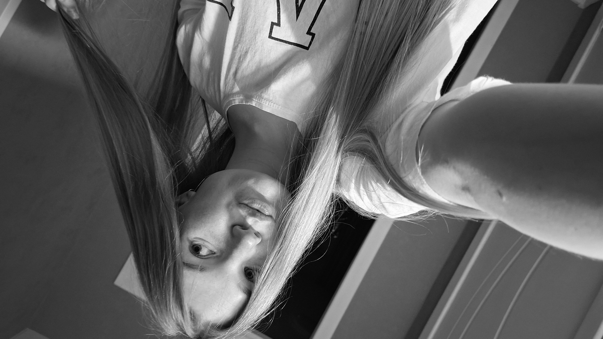 A young Caucasian female with long, straight blonde hair is shown in a close-up, black and white selfie. She is wearing a white t-shirt with a large black letter "K" visible on the right side of the chest. Her head is tilted, and her hair is draped across her face and body. She is looking directly at the camera with a slight pout. The background appears to be indoors, with horizontal lines suggesting blinds or a similar textured surface. There are shadows on her arm and t-shirt, indicating directional lighting. The image is slightly distorted due to the angle of the selfie. No specific location in Chaikovsky, Russia, can be identified from the image.