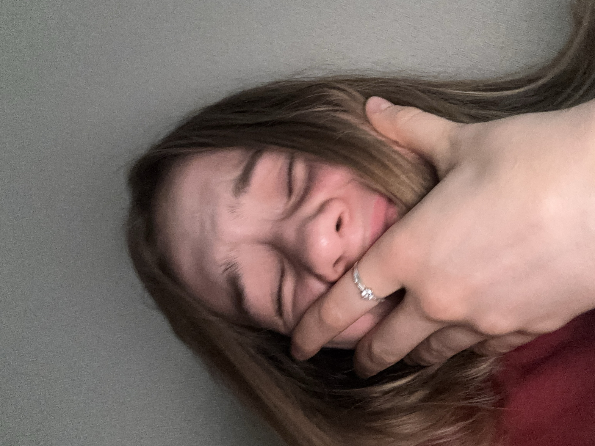 This image is a close-up, self-taken photograph of a young person with long, light brown hair. Their eyes are closed, and their face appears distressed, with their mouth open and their hand covering part of their mouth and nose. They are wearing a ring with a small stone on their right index finger and a red garment, possibly a shirt or sweater, on their left side. The background is a plain, textured gray wall. There is no information in the image to suggest a location in Vsevolozhsk, Russia.
