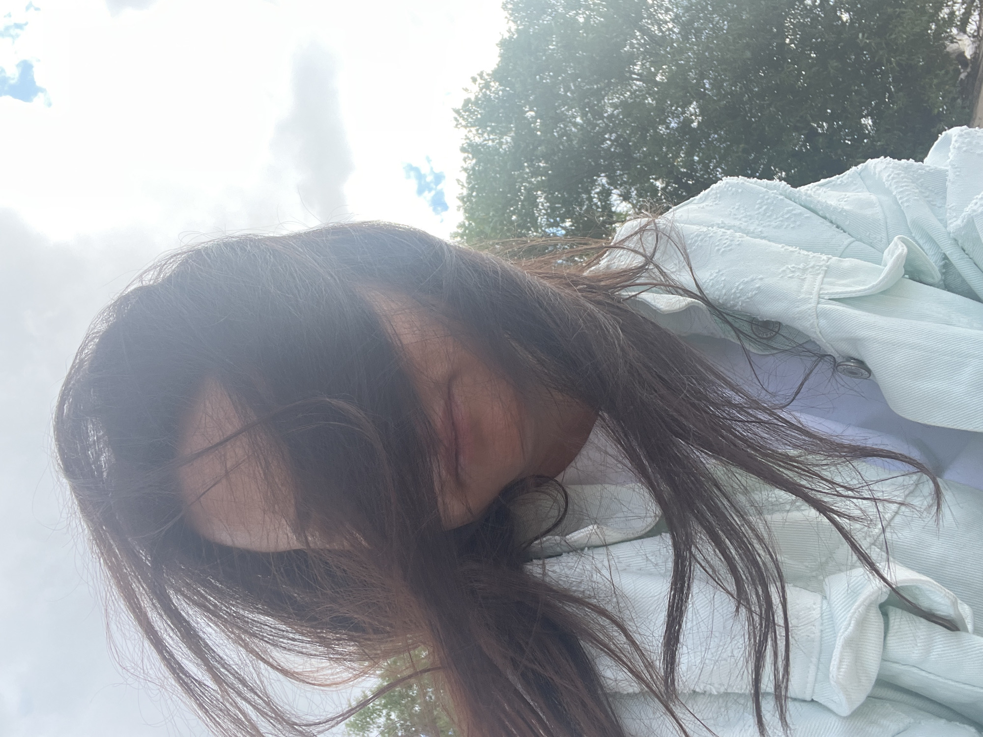 A medium shot, taken from a low angle outdoors on a cloudy day, shows a person with long, dark hair blowing across their face. The person is wearing a light blue textured jacket over a white t-shirt. Their mouth is visible, but their eyes are obscured by their hair. In the background, trees and sky are visible. The specific location in Saint Petersburg, Russia cannot be determined from the image.