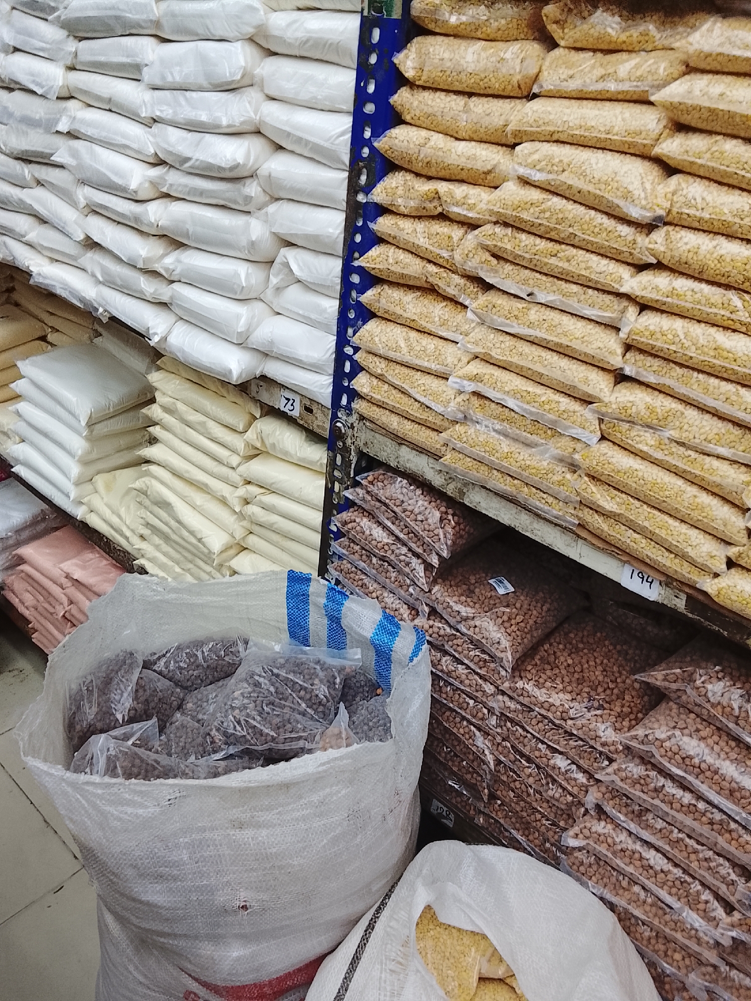 The image depicts a store interior in Gulgasht Colony, Pakistan. It shows shelves laden with sacks and plastic bags filled with various food staples, likely grains, beans, or flour. The topmost shelves are stacked with white sacks, possibly flour or rice. Below these are rows of clear plastic bags containing yellowish granular material, potentially lentils or other pulses. Another shelf holds bags of brown beans or chickpeas. A large open sack in the foreground contains smaller bags of dark-colored beans. The scene suggests a wholesale or bulk food market.
