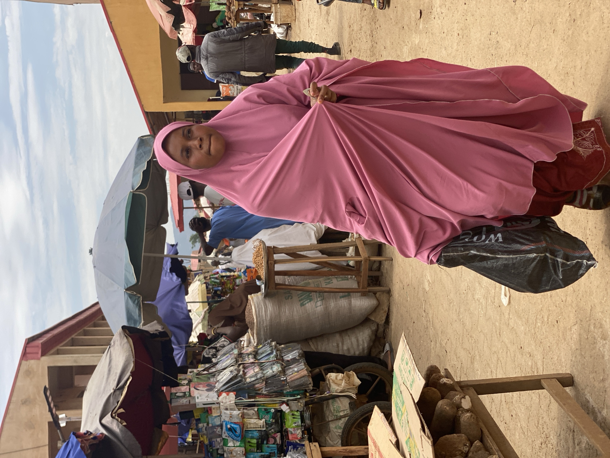 The image depicts a woman wearing a pink hijab in a bustling market setting, likely in the city of Kafanchan B, Nigeria. She is holding the fabric of her hijab in her hand. The market displays a variety of goods, including electronics, food items, and bags of what appears to be grain or beans, stacked on carts and tables. The background features other individuals, some under umbrellas or canopies, and buildings.