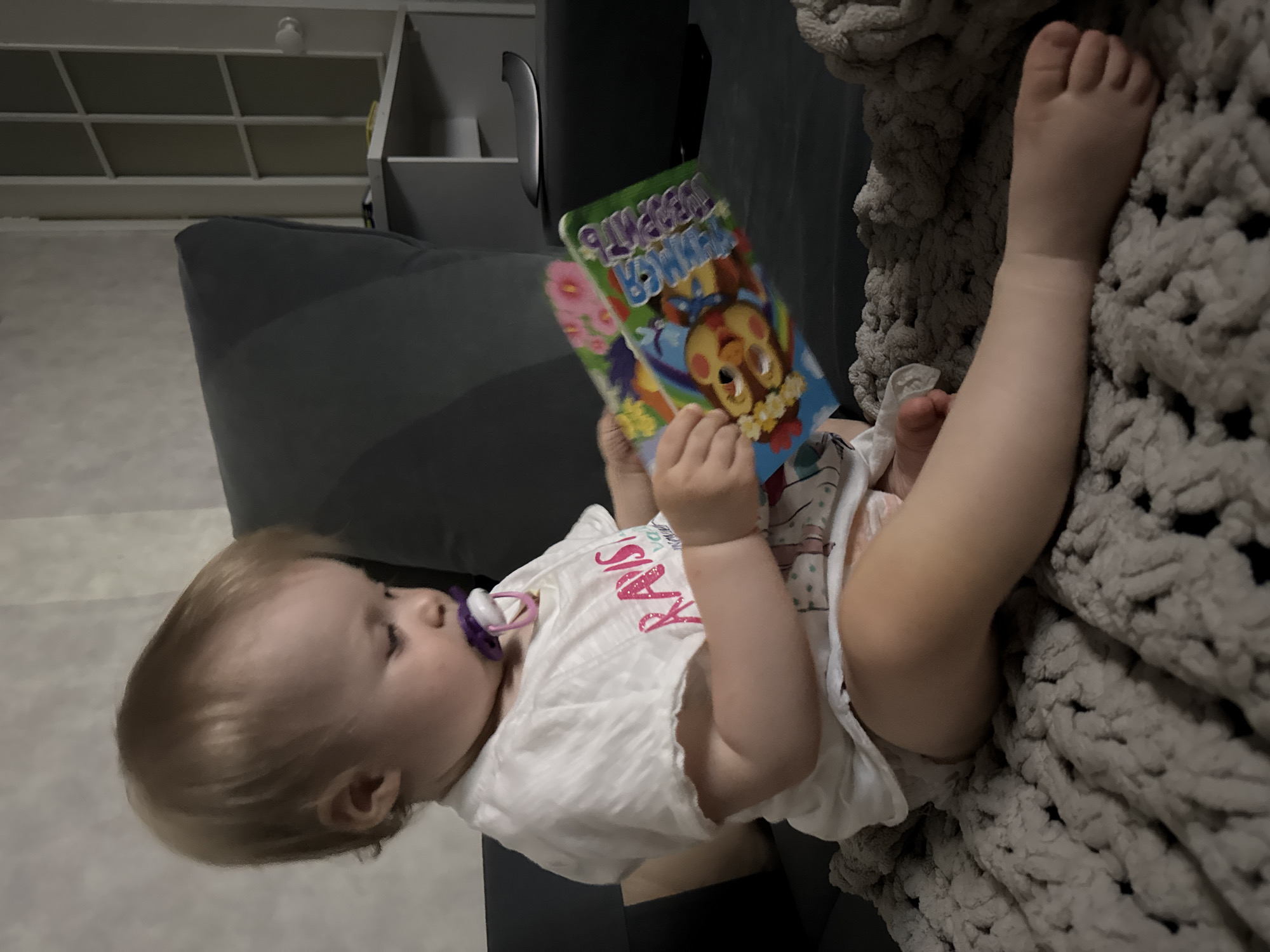 A baby with blond hair is reclining on a gray sofa. The baby is wearing a white T-shirt and a diaper. The baby is holding a brightly colored children's book in their hands and has a pacifier in their mouth. The baby's legs are extended and resting on a thick, gray, textured blanket. The background includes a built-in shelf unit with white shelves and a gray cushioned surface. The location is unspecified.