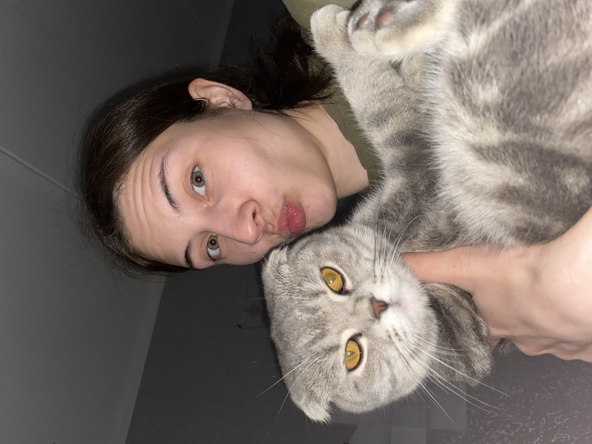 A woman with dark hair pulled back, fair skin, and green eyes is posing in a selfie with a gray and white tabby cat. The woman is puckering her lips slightly towards the camera. The cat has yellow eyes and folded ears, likely a Scottish Fold. The woman is holding the cat close to her face, and the cat's body is oriented towards the upper right corner of the frame. The background appears to be a dimly lit room with light gray walls. The location is not discernible from the image and cannot be identified as Votkinsk, Russia.