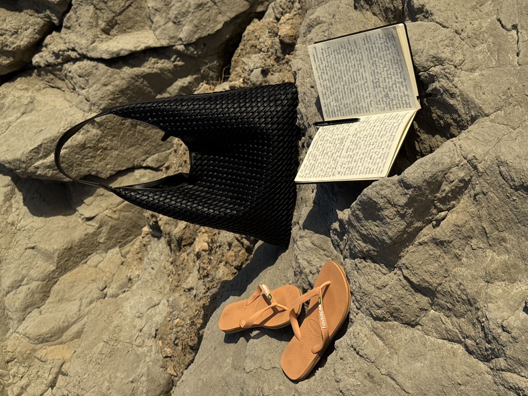 A black woven tote bag, a pair of orange flip-flops, and an open notebook with a pen are arranged on a rocky surface. The tote bag is on the right, the flip-flops are in the lower left, and the notebook is in the center. The scene appears to be outdoors, possibly near a beach, given the rocky terrain. The lighting suggests it is daytime, with sunlight casting shadows. The location is not Biarritz, France, as no visual cues indicate this specific city.