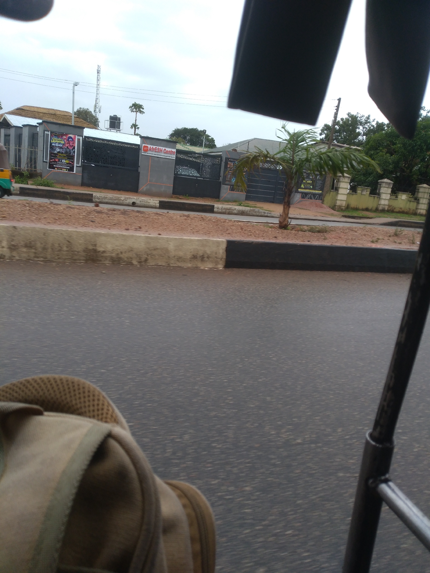 The image presents an outdoor street scene, likely captured from a vehicle given the perspective and visible internal parts in the foreground. In the bottom left, a section of a light brown bag with a mesh backpad is visible, while a dark metal bar, part of a vehicle frame, cuts into the frame from the right.
The mid-ground features a wet, asphalt road, suggesting recent rainfall. It is bordered by a concrete curb with black and white sections, and a raised median of reddish-brown dirt or gravel. Beyond this, a black fence with decorative cutouts and patterns encloses a property.
On the fence, two prominent signs are displayed. To the left, a large white poster advertises "SLAUGHTER HOUSE Mr RaNgeR" with additional text like "DANCE HALL," "AFRICAN," and "REGGAE," alongside images of two men. To its right, a red and white sign reads "AfriESH Centre" with a tagline indicating it provides "multi-vendor data & cyber-security solutions." A small palm tree stands near the fence.
The background shows various buildings, one with a dark roof and another with a thatched roof. A tall cellular tower and a street light pole are visible against an overcast, grey sky. Lush green trees and foliage are scattered across the scene. A small, partially visible green and yellow auto-rickshaw is on the far left. The scene depicts a quiet, potentially urban or semi-urban area, under cloudy conditions.