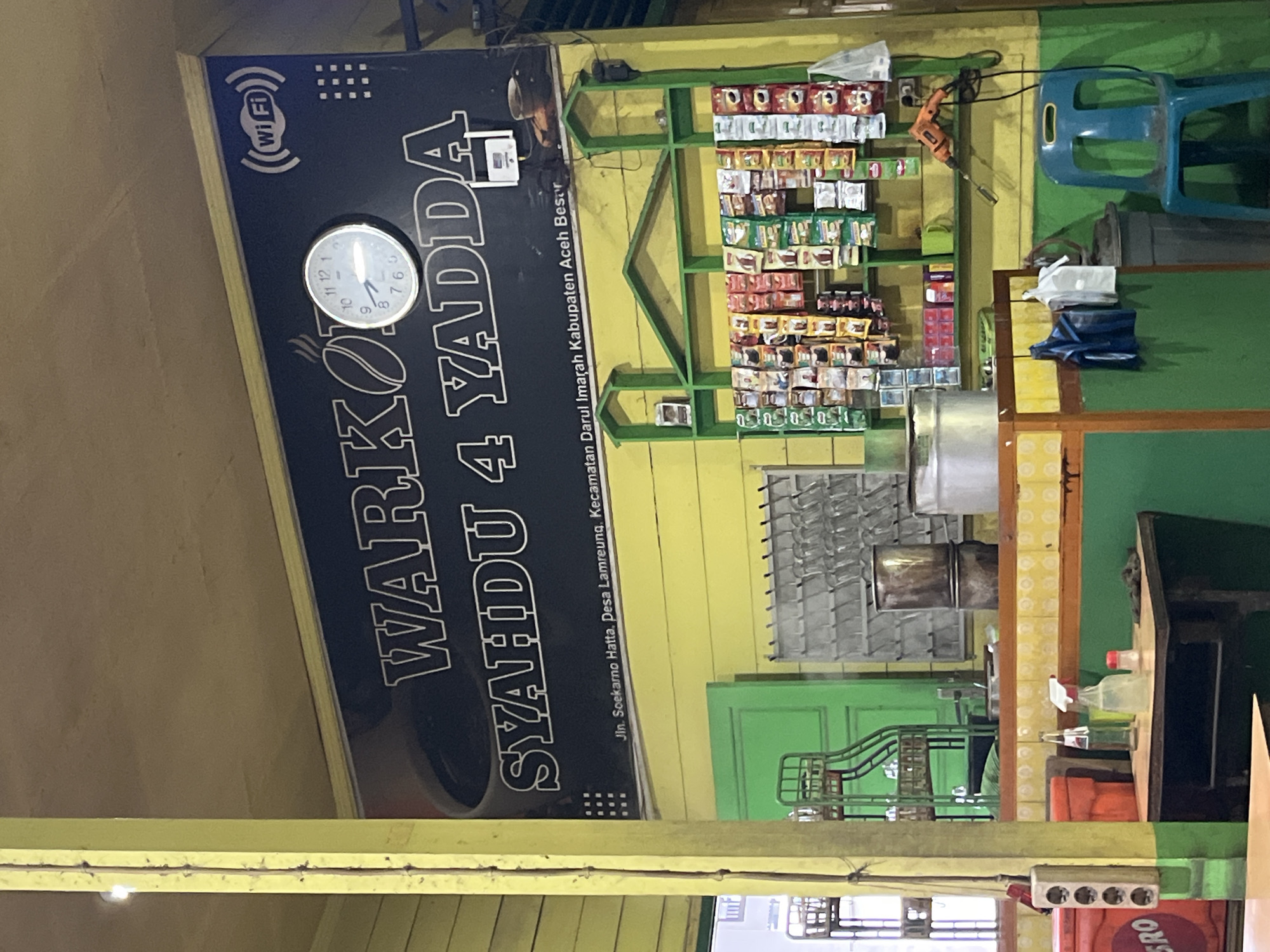 The image depicts the interior of a "Warkop Syahdu 4 Yadda" in Lampeuneurutblang, Aceh Besar, Indonesia. The warkop, or coffee shop, is characterized by its simple, rustic interior. A large black sign dominates the upper portion of the wall, displaying the warkop's name in white lettering. To the right of the name, a round analog clock with black hands shows the time as approximately 9:50. Above the clock, a white "Wi-Fi" logo is visible. Below the warkop's name, smaller text indicates the address: "Jln. Soekarno Hatta, Desa Lamreung, Kecamatan Darul Imarah, Kabupaten Aceh Besar".
The walls of the warkop are painted yellow and green. A green shelf unit is mounted on the yellow wall, displaying numerous packets of snacks and instant food items. To the left of the shelves, a green metal rack holds what appear to be glass cups. In the lower section of the image, a counter area is visible, with various items on top, including bottles, a spray bottle, a salt shaker, and a cloth. Behind the counter, a large metal container sits on the counter. To the right of this, a blue plastic chair is positioned next to a grey trash can. Electrical outlets are mounted on a yellow pole on the left side of the image. The overall impression is of a casual, local gathering spot.