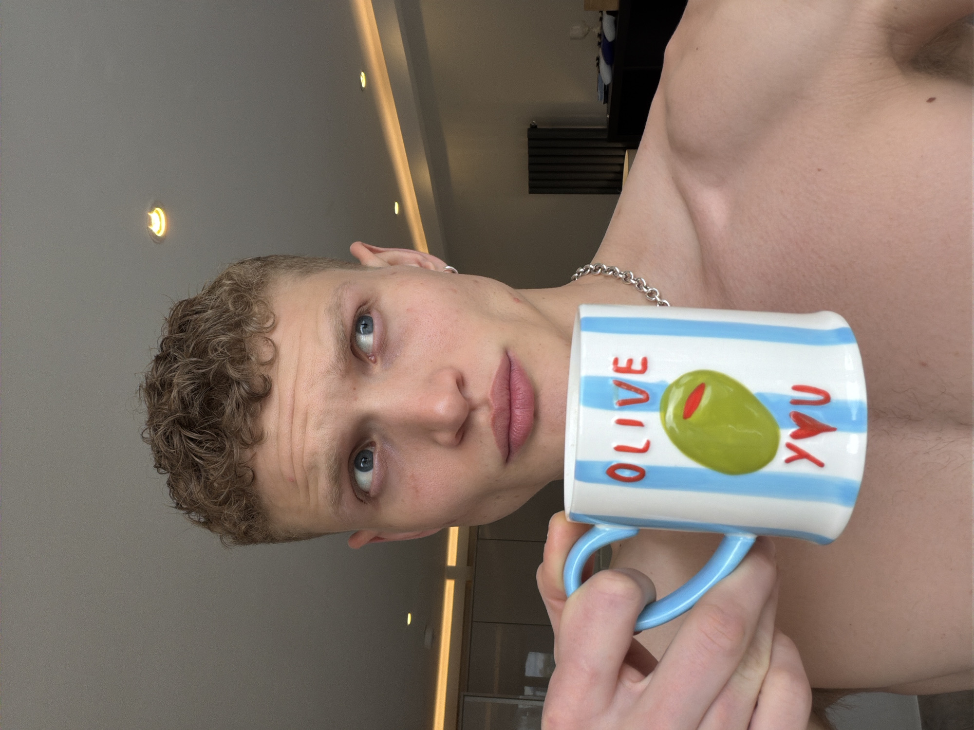 A close-up, indoor portrait shows a fair-skinned person with short, light brown curly hair and blue eyes holding a mug. The mug is white with blue horizontal stripes and features the words "OLIVE" and "YOU" in red, with a green olive graphic between them. The person is looking directly at the camera with a neutral expression, showing their lips and nose. The person is wearing a silver chain around their neck. Their chest and shoulders are visible, suggesting a casual or shirtless state. The background includes a light-colored wall with recessed lighting and glimpses of a modern interior, possibly a kitchen or living area with cabinets. The location is not identifiable as being in the city of London, United Kingdom.