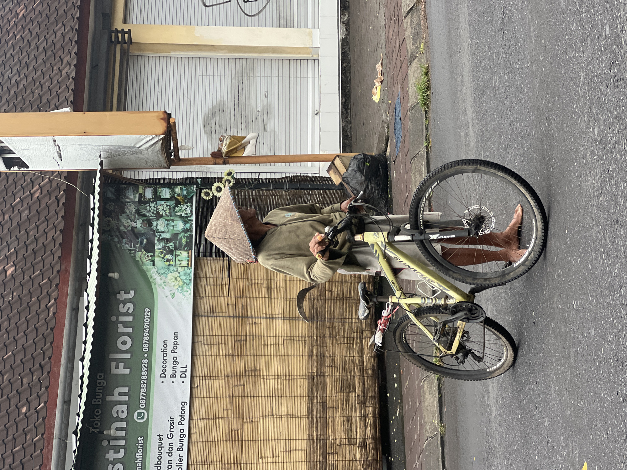 A medium, outdoor shot depicts an Asian man leaning against a roadside structure, with a bicycle positioned in front of him in Sukawati, Indonesia. The man wears a traditional conical hat and an olive-green long-sleeved shirt and trousers. He appears to be engaged in a cell phone. His bare feet rest on the street surface.
A light green mountain bike, brand name "Atlantis," stands on the right side of the frame, with its front wheel closer to the viewer. A black plastic bag is adjacent to the man.
Behind the man is a banner for "Stihah Florist," featuring a WhatsApp logo and a phone number, along with bullet points indicating services like decoration and bunga papan (flower arrangement in Indonesian). A bamboo-screen wall is also visible. The storefront’s metal roll-up door is closed. The street pavement occupies the lower right of the frame.
