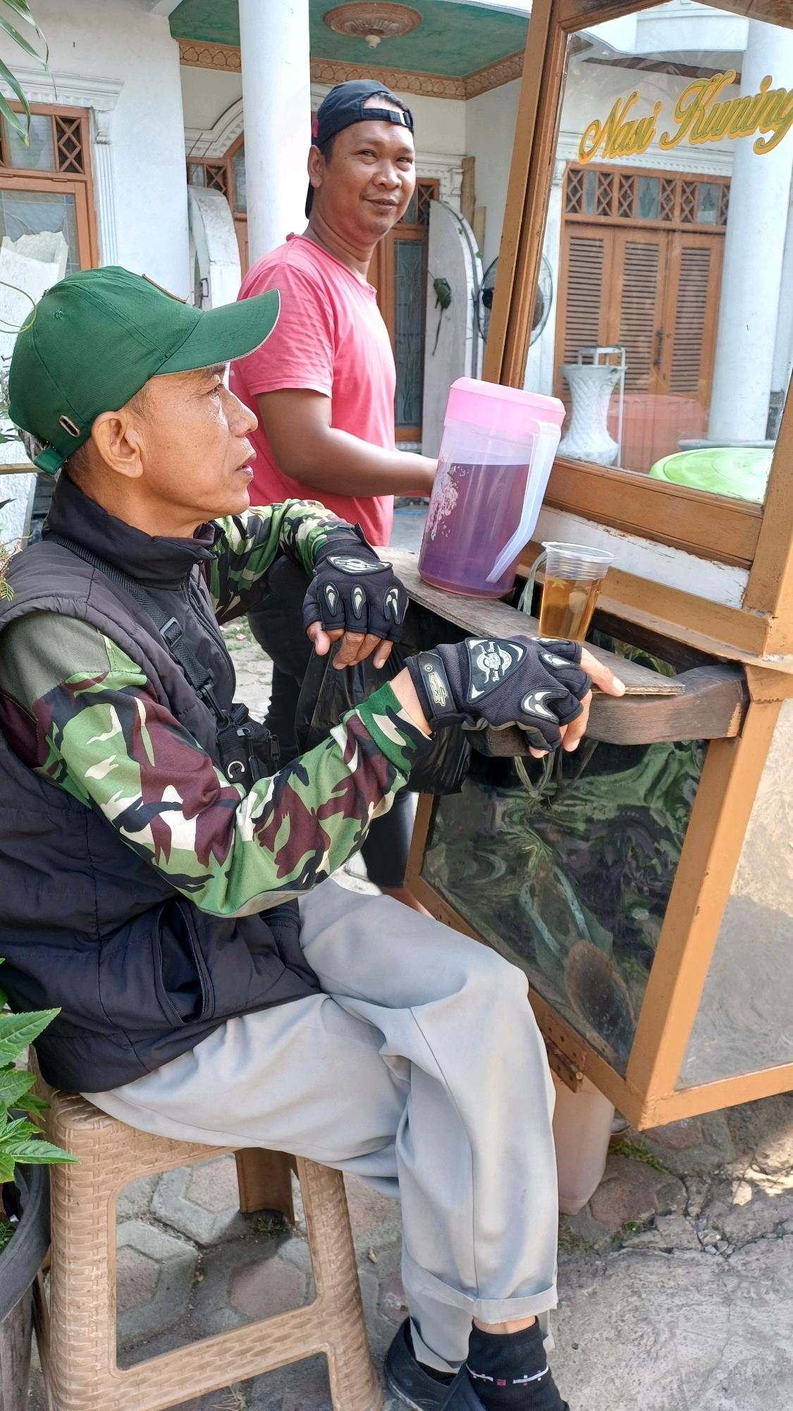 In Gunung Putri, Indonesia, two men are present. The man in the foreground, wearing a green cap, a black vest over a camouflage shirt, and light gray pants, sits on a tan plastic stool. He is looking to his left. The second man, standing behind him and to his right, wears a red t-shirt and a black baseball cap, looking towards the camera. He is holding a large pink plastic jug with purple liquid. In front of them is a wooden food cart with glass panels, displaying what appears to be food items within. A clear plastic cup with a light brown liquid is on the cart's wooden surface. The background shows a building with white pillars and wooden-framed windows, along with a portion of a green roof. The ground is paved with hexagonal bricks and some scattered dirt.