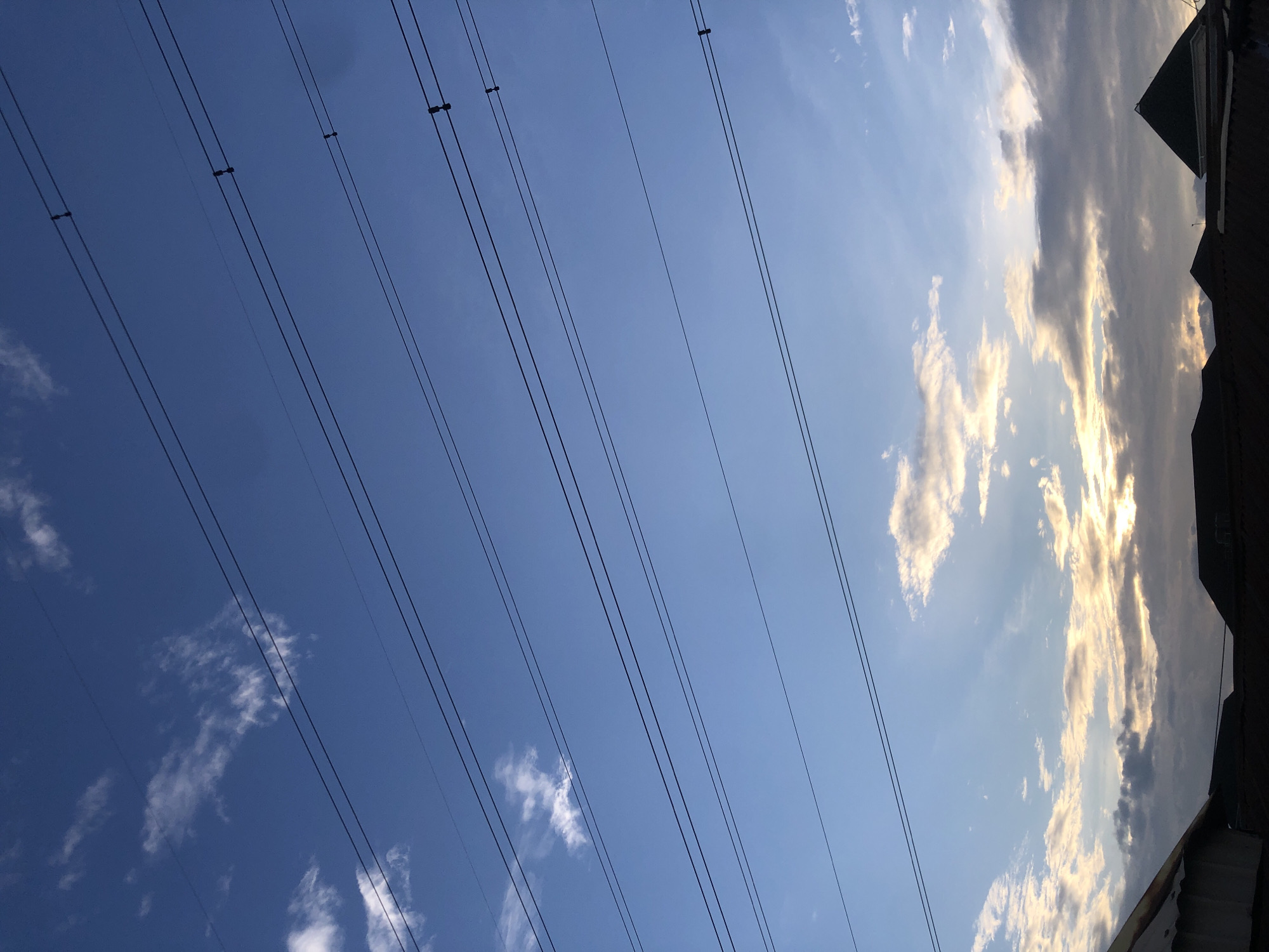 The image shows a clear blue sky with scattered white clouds. Multiple power lines stretch horizontally across the frame, supported by insulators. In the lower part of the image, the silhouettes of rooftops of houses are visible, suggesting an urban or suburban environment. The lighting indicates it might be late afternoon or early morning, with the sun low on the horizon, casting a warm glow on some of the clouds. The scene is devoid of people or any specific activity. The perspective suggests the photo was taken from a slightly elevated position, looking upwards at the sky and downwards at the rooftops. The presence of power lines is a common feature in many urban and suburban landscapes in cities like Ado, Nigeria, where infrastructure for electricity distribution is prevalent.