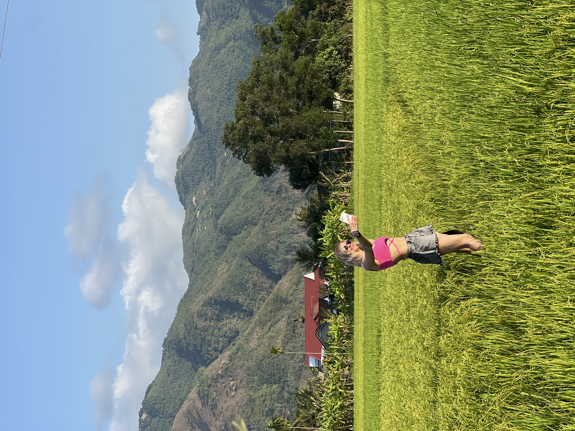 A woman is standing in a rice field in Diên Khánh, Vietnam. She is wearing a pink top and jean shorts and is holding a smartphone in her hand, apparently taking a selfie. The rice field is green and extends into the distance. In the background, there are mountains covered with trees. There is also a building with a red roof partially visible. The sky is blue with some white clouds.