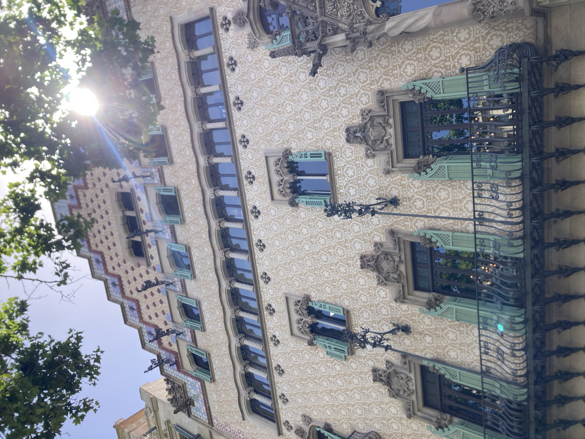 The image shows the facade of Casa Batlló, a building in Barcelona, Spain, designed by Antoni Gaudí. The building's exterior is adorned with intricate details, including a mosaic of broken ceramic tiles and curved balconies that resemble bones. The image is taken from a low angle, looking up at the building. The sun is shining brightly, casting a lens flare over the top of the image. Green foliage from trees can be seen in the foreground, partially obscuring the building. The architecture is characterized by its organic forms, vibrant colors, and playful details, reflecting Gaudí's unique Art Nouveau style.